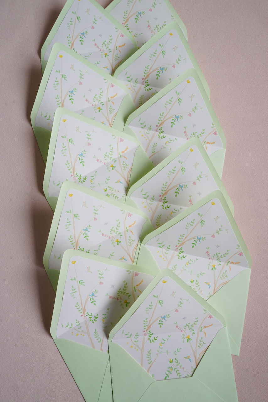 A group of light green envelopes partially open, each containing a card with a delicate, pastel-colored tree branch design with small leaves and tiny flowers or birds. The envelopes are arranged in a slightly overlapping, cascading pattern on a pink surface.