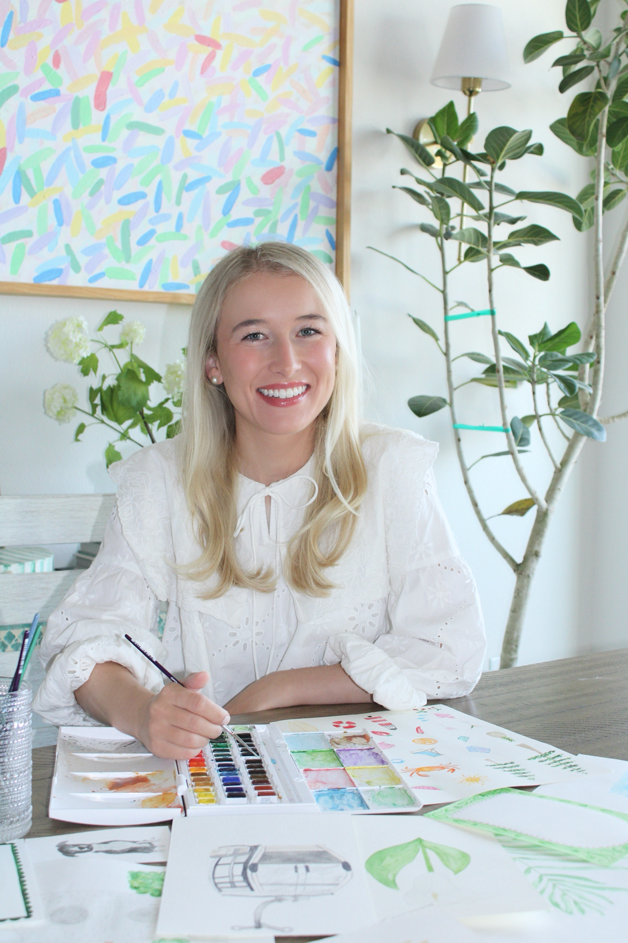 A woman with long blonde hair smiling and seated at a wooden table, holding a paintbrush, surrounded by watercolor paintings and art supplies, in a room with greenery and abstract artwork on the wall.