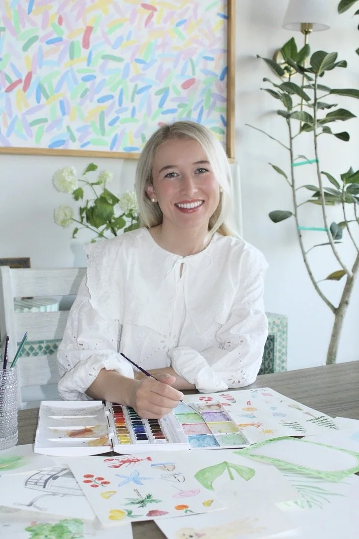 A young woman with blonde hair and a white blouse sitting at a table with watercolor paints and drawings, smiling at the camera, in a bright room with plants and colorful artwork in the background.