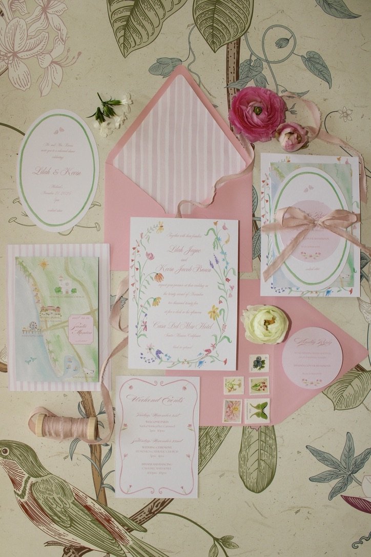 Open pink and white floral wedding invitation suite with matching envelope, small decorative stamps, and pink flowers on floral-patterned background.