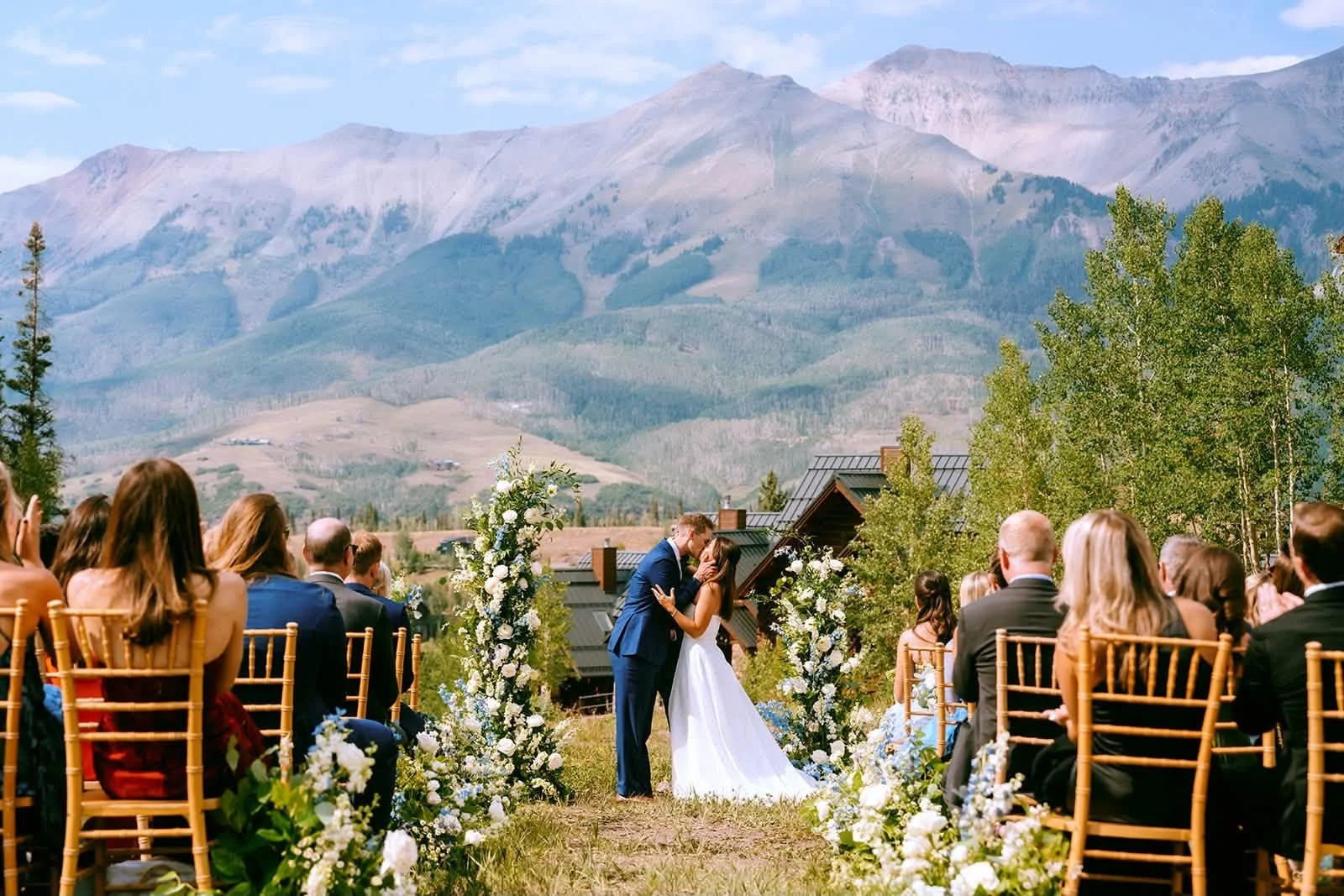 A wedding ceremony outdoors with a mountainous landscape in the background. A bride and groom stand in the center, kissing, surrounded by floral arrangements, with guests seated on either side.