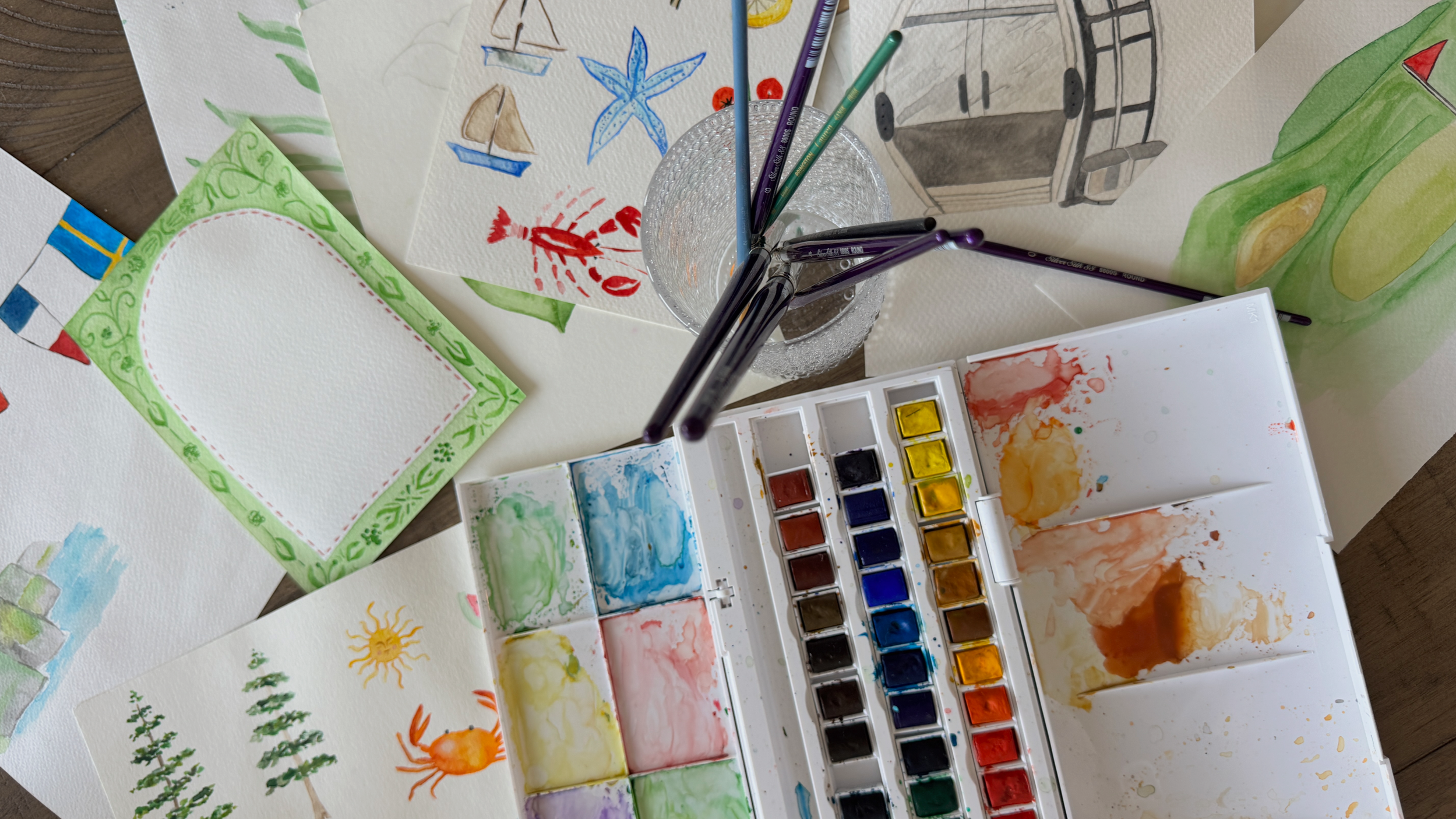 Watercolor paintings, art supplies, and a glass holding brushes on a wooden table.