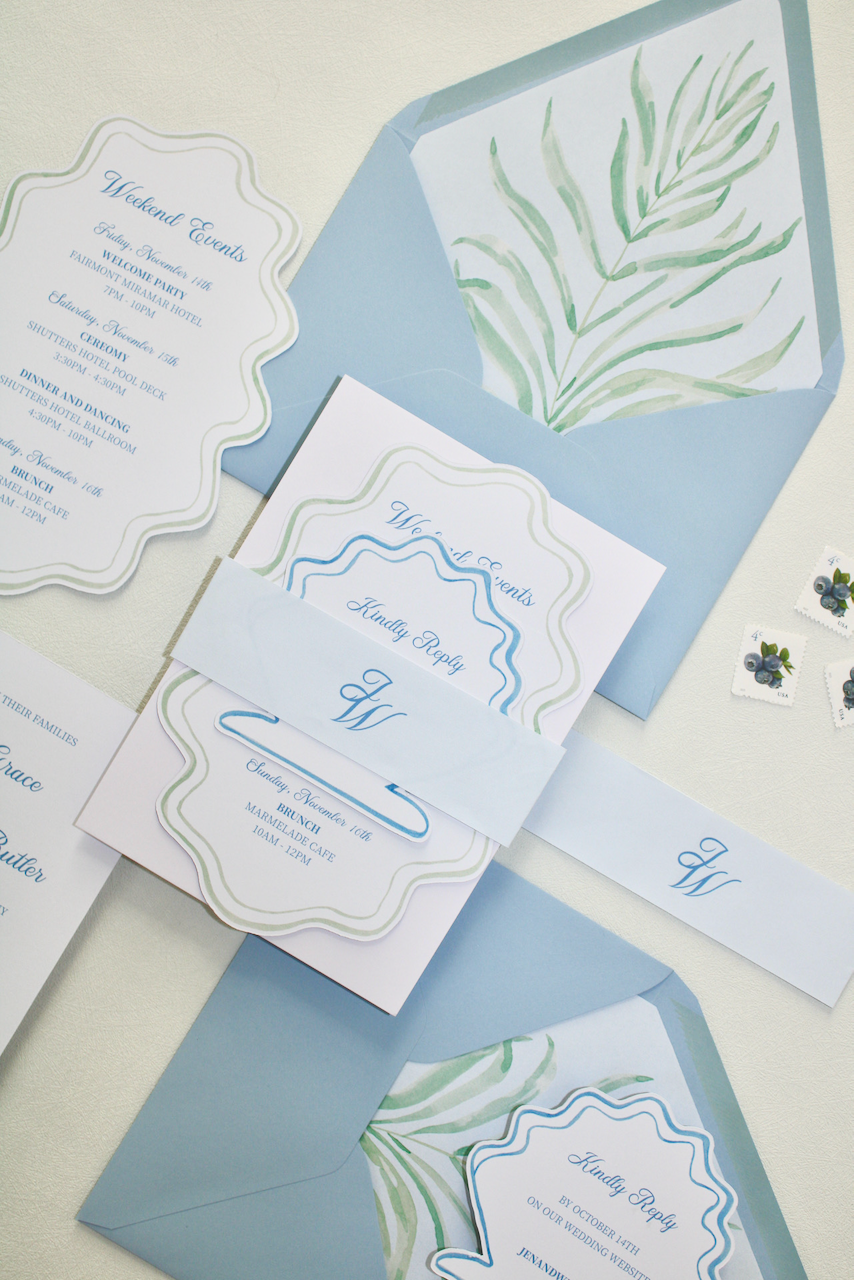 Wedding invitation suite with matching envelopes, reply card, and event details, featuring watercolor green foliage and blue accents.