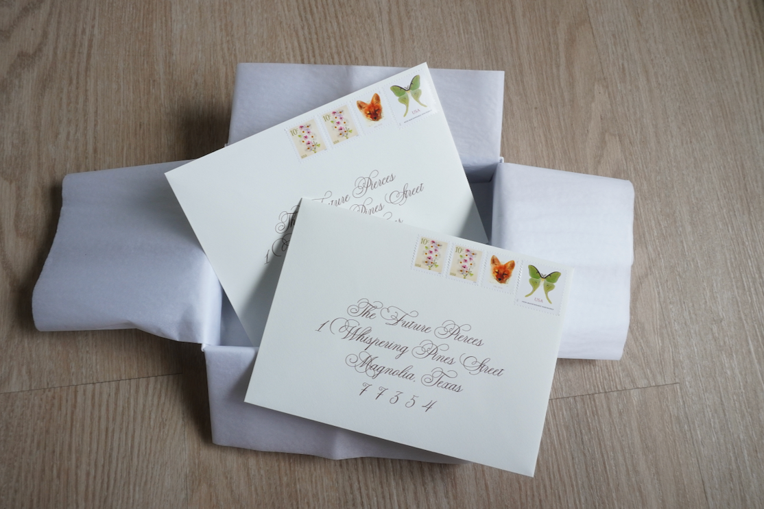 Two white envelopes with decorative postage stamps, placed on a wooden surface with white tissue paper