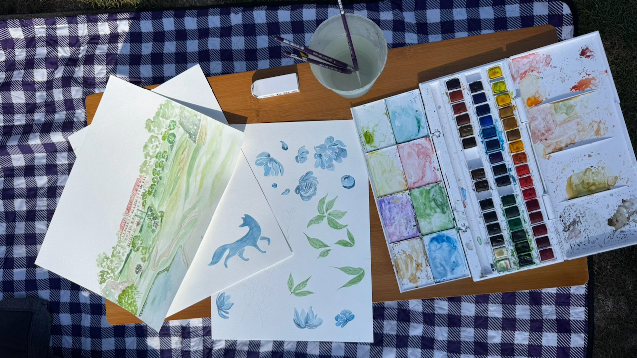 Watercolor painting supplies on a table, including a watercolor palette, paper with painted sketches of plants and animals, and a mug with brushes, outdoors on a checkered tablecloth.