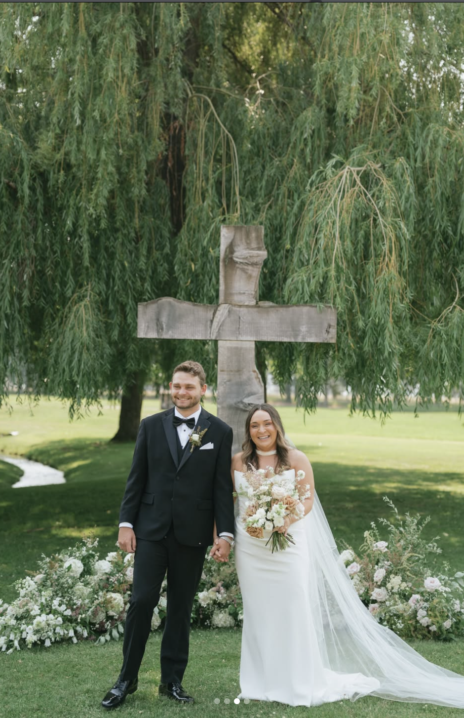 A newlywed couple standing outdoors in front of a wooden cross and lush green tree, holding hands and smiling, with floral arrangements at their feet.