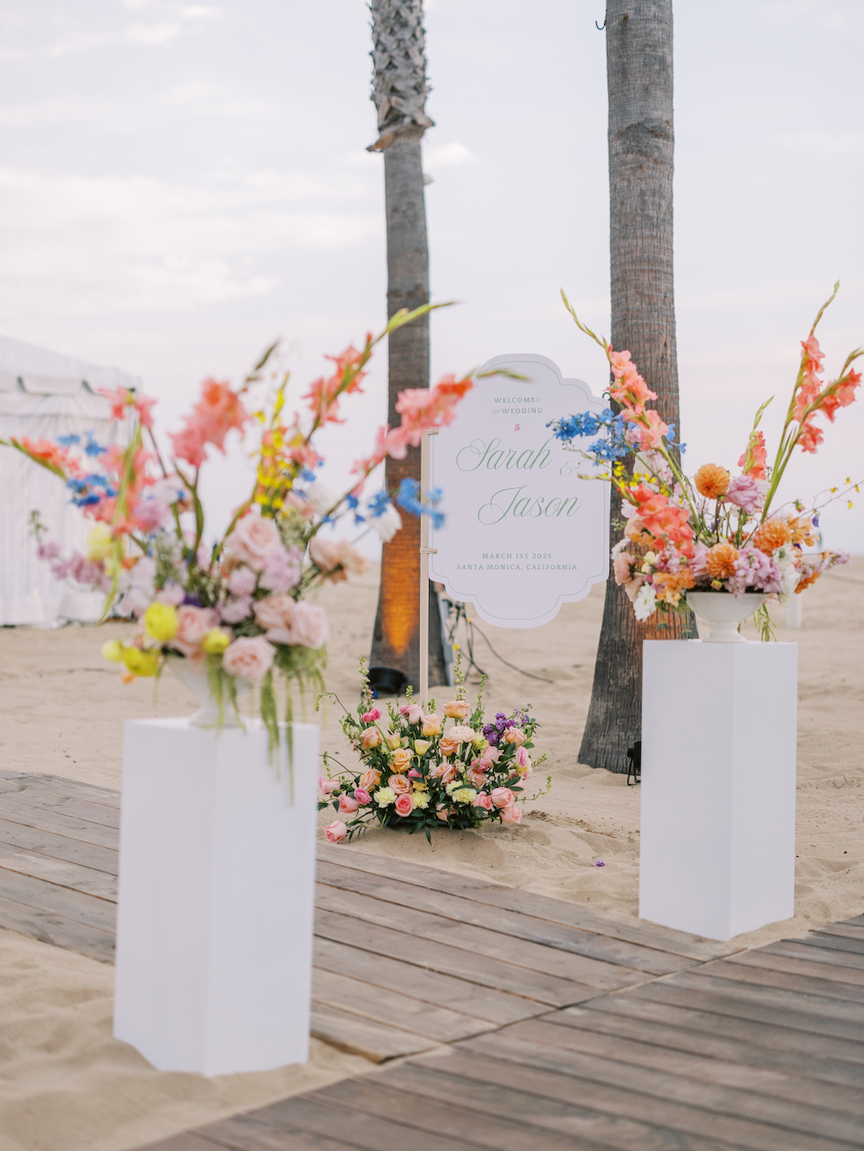 Wedding decoration on a beach with floral arrangements and a sign for Sarah and Jason's wedding on March 1st, 2025, in Santa Monica, California.