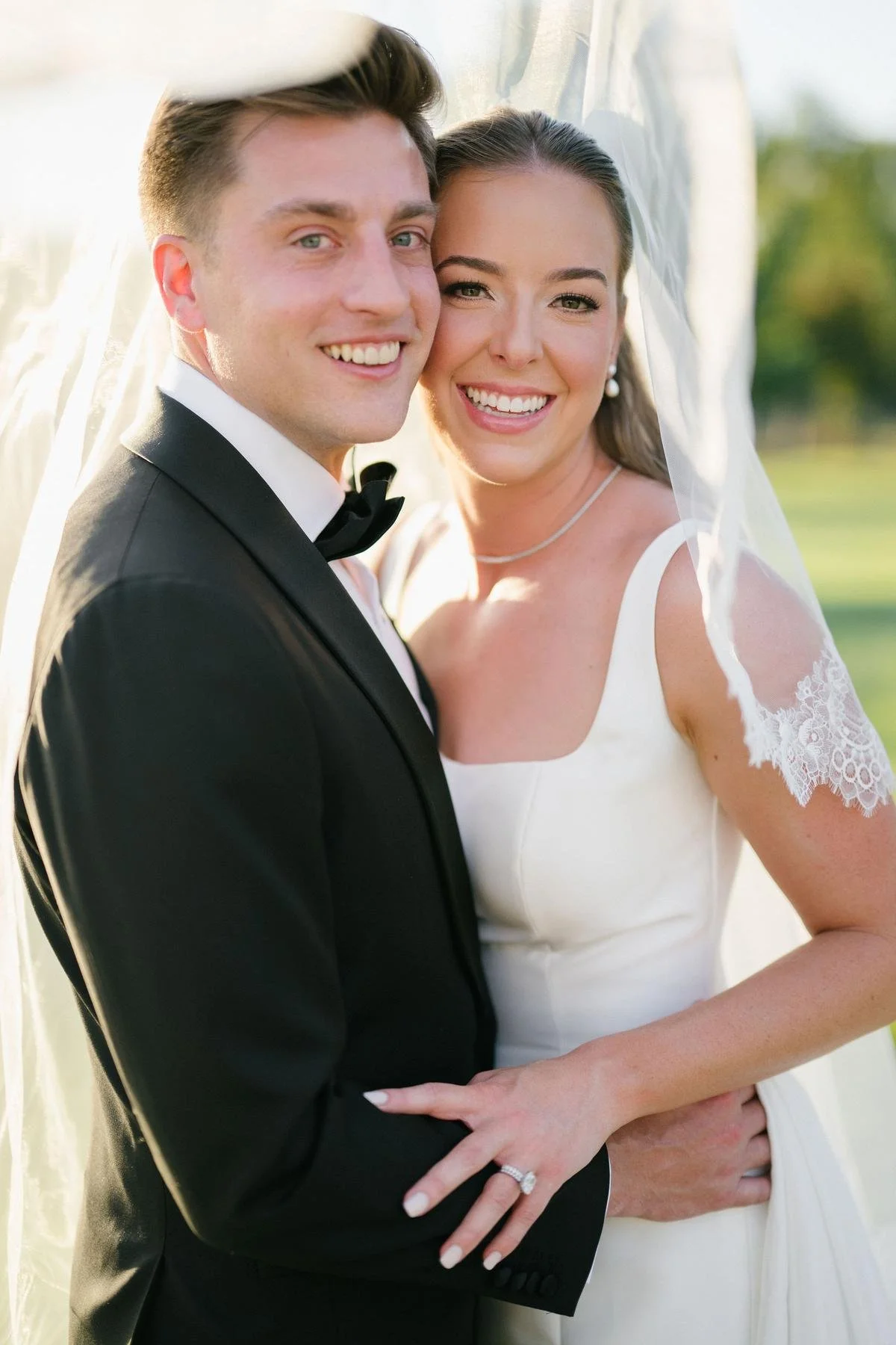 A newlywed couple smiling and embracing outdoors, dressed in wedding attire with the groom in a tuxedo and the bride in a white wedding gown and veil.