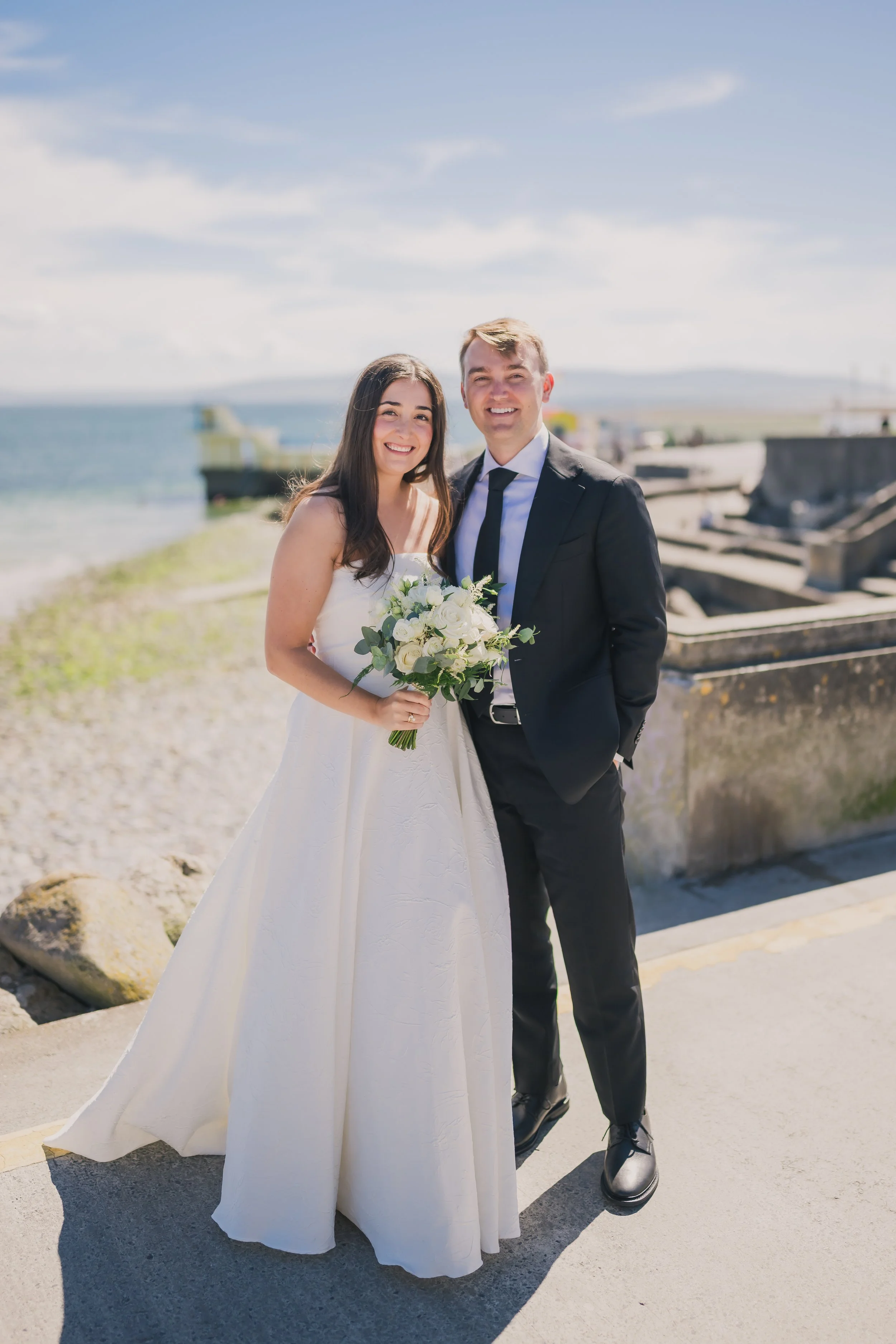 A bride and groom smiling on a beach, with the bride holding a bouquet of flowers, and the groom in a black suit. The sky is partly cloudy, and there are rocks and a boat in the background.