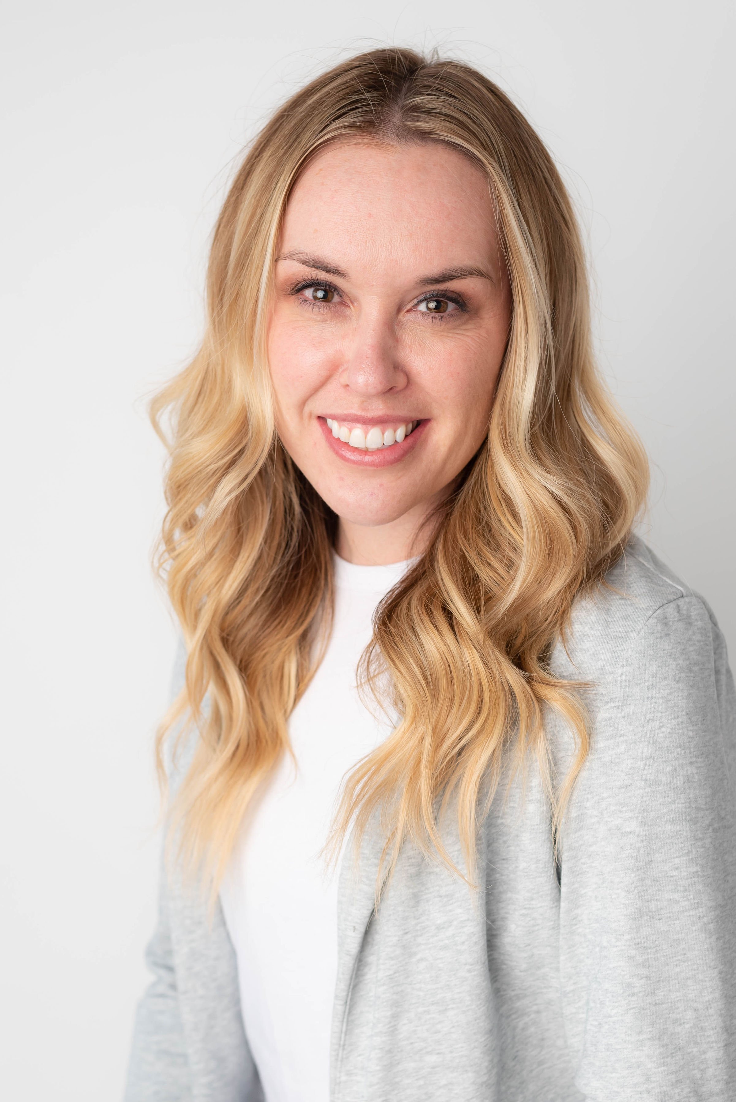 A woman with long wavy blonde hair smiling and looking directly at the camera, wearing a light gray zip-up hoodie over a white shirt, against a plain light background.