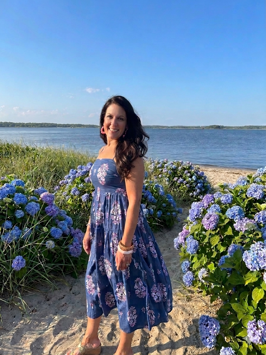 Woman smiling on a beach surrounded by blue and purple hydrangea flowers, wearing a blue floral dress with a waterfront in the background under a clear blue sky.