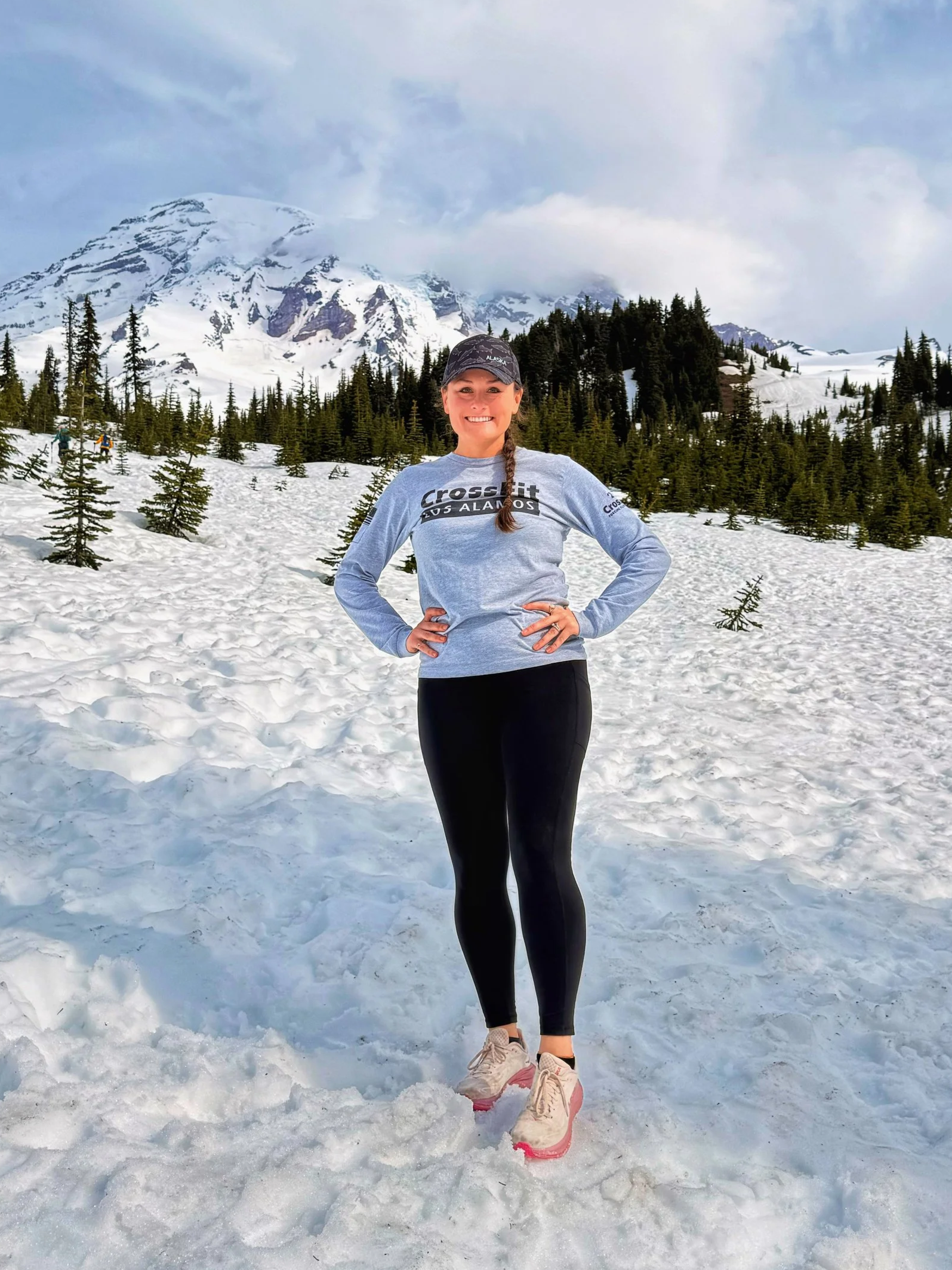 A woman standing on snowy ground in front of snow-covered mountains and pine trees, wearing athletic clothing and smiling at the camera.