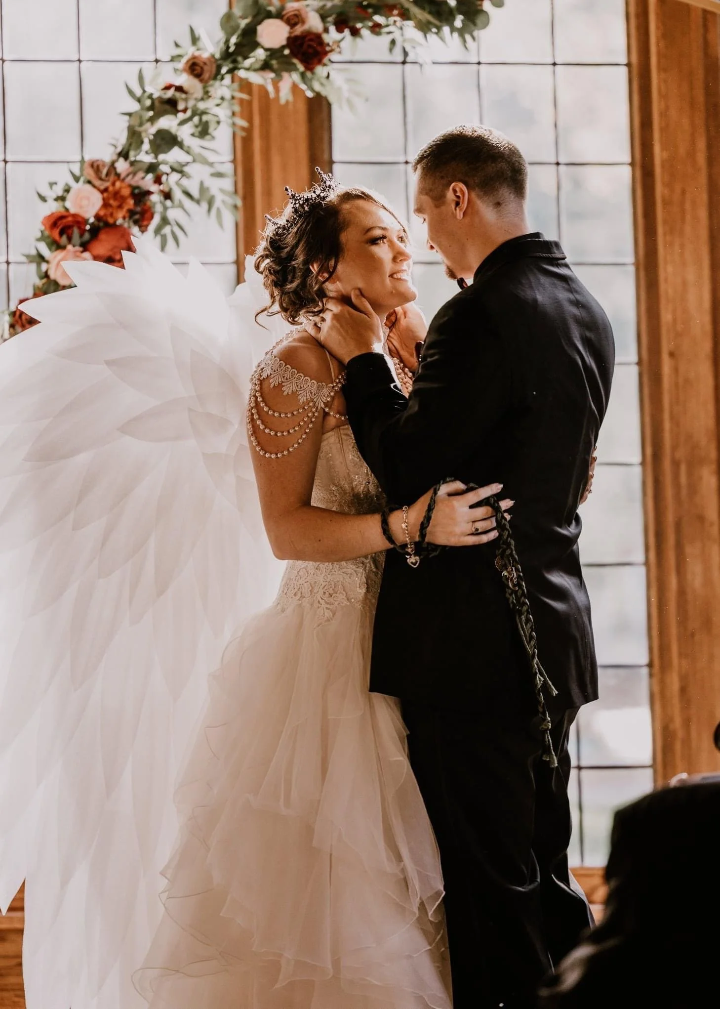 A bride and groom during their wedding ceremony, standing close and gazing at each other affectionately, with a floral arch in the background and a wooden wall behind them.