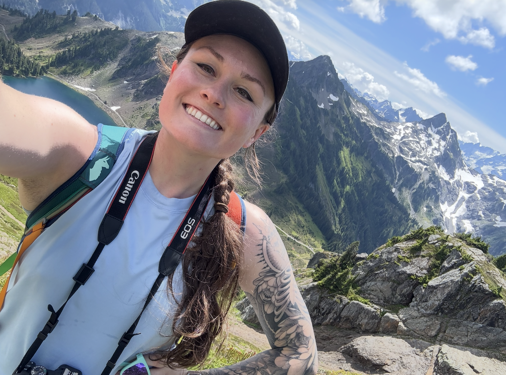 A smiling woman taking a selfie outdoors in a mountainous landscape with snow-capped peaks, rocky terrain, green vegetation, and a lake in the background. She is wearing a black cap, a sleeveless white top, and has a camera strap around her neck, with tattoos visible on her arm.