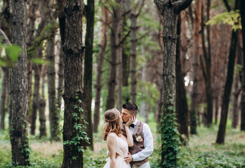 A couple kissing in a lush green forest with tall trees and dense foliage.
