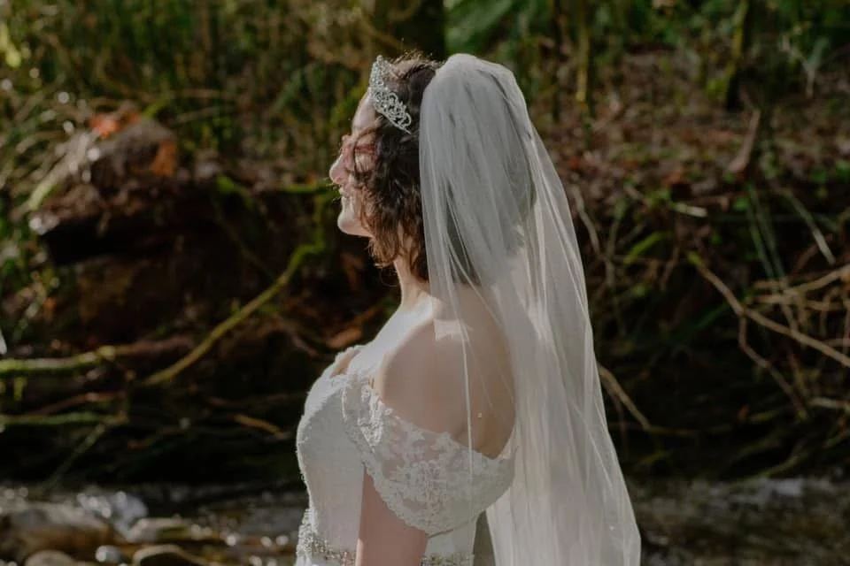 A woman dressed in a wedding gown with a lace off-shoulder neckline and a bridal veil is outdoors in a natural setting with greenery and a stream in the background.