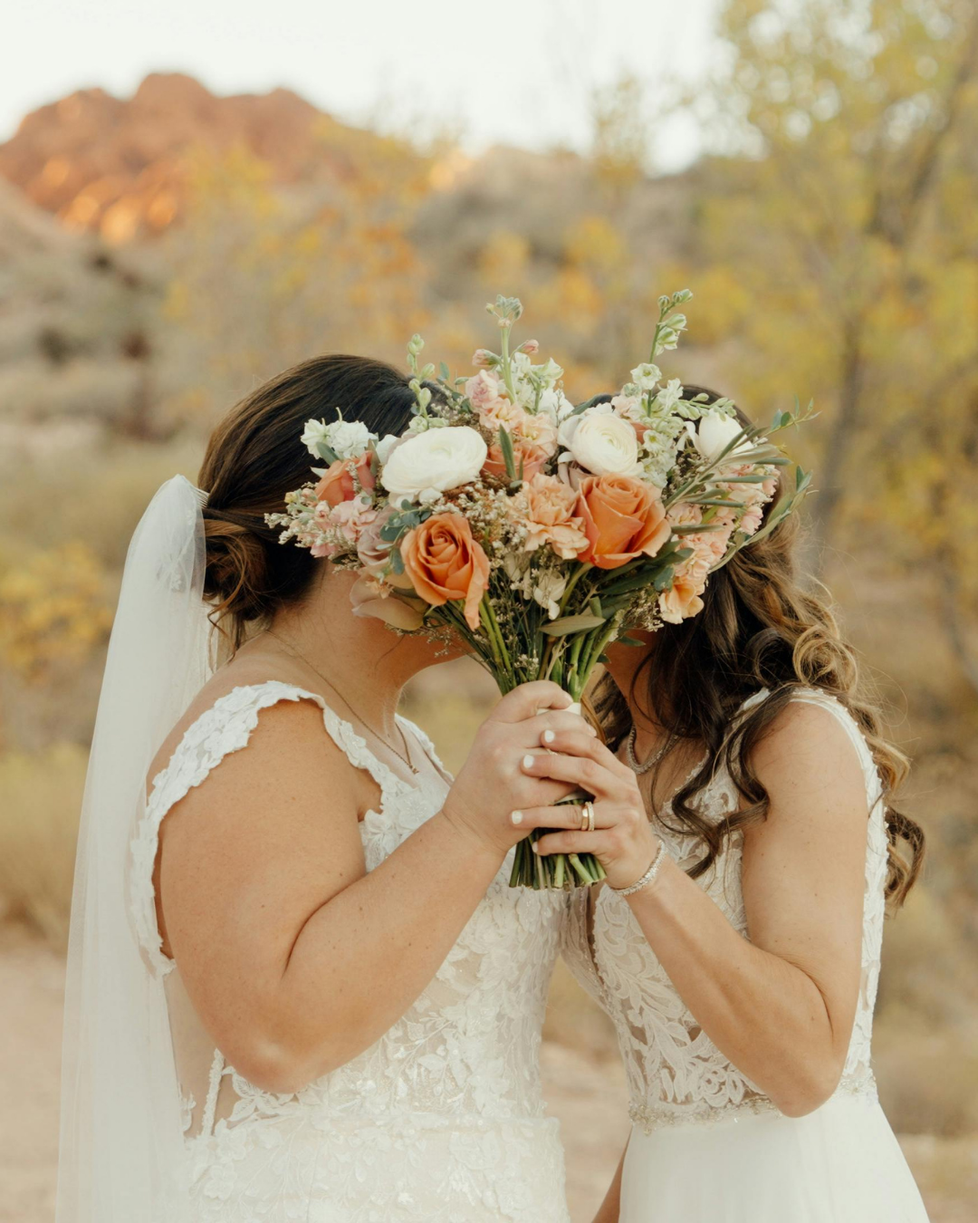 Two women in wedding dresses holding a bouquet of flowers, one with dark hair and the other with wavy hair, outdoors with trees and rocky hills in the background.