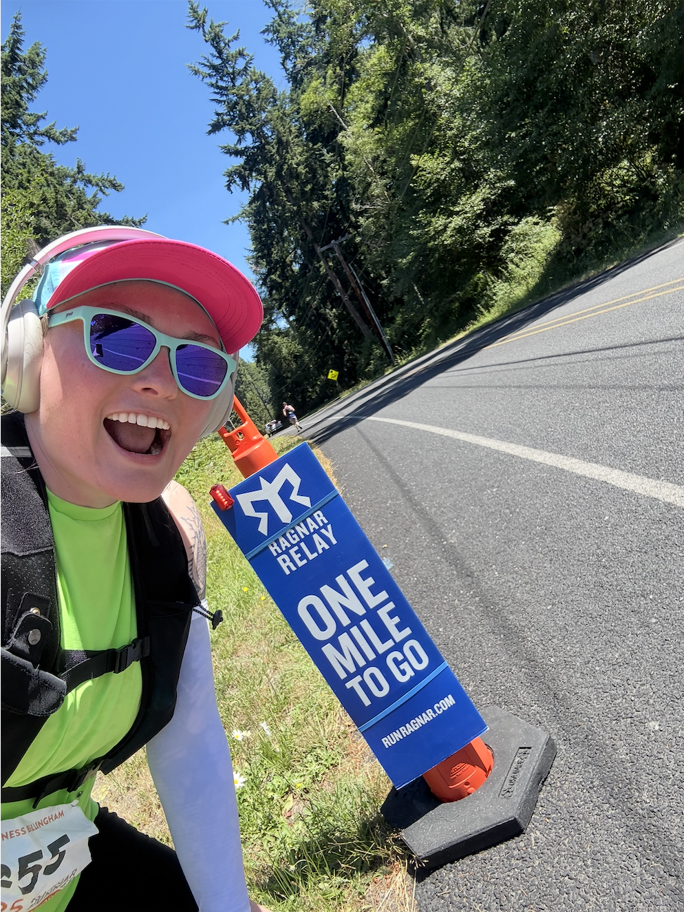 A joyful woman with sunglasses, a pink cap, and headphones, smiling next to a sign that reads 'Ragnar Relay One Mile to Go' on a roadside with trees in the background.