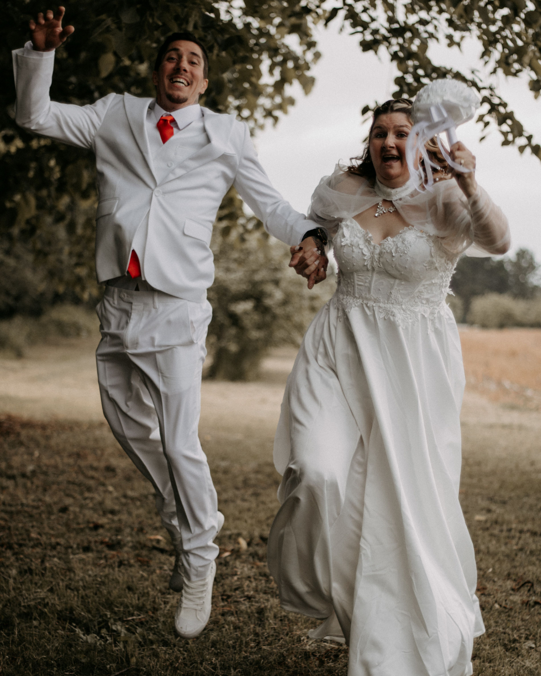 A bride and groom holding hands and running outdoors, the bride is holding a white wedding bouquet, and they both appear joyful and excited.
