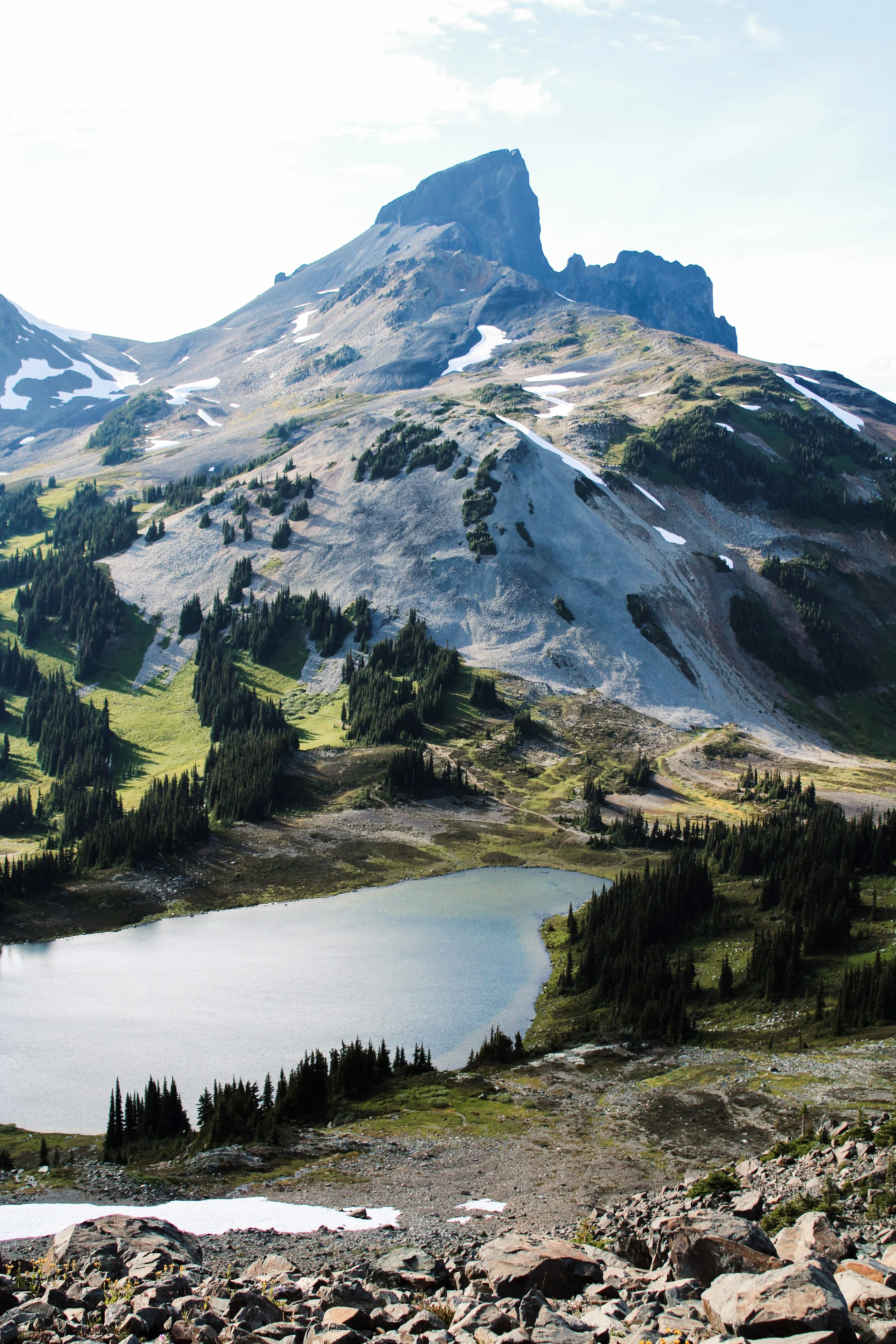 A mountainous landscape featuring a tall rocky peak, patches of snow, green forests, a lake at the base, and rocky terrain in the foreground.