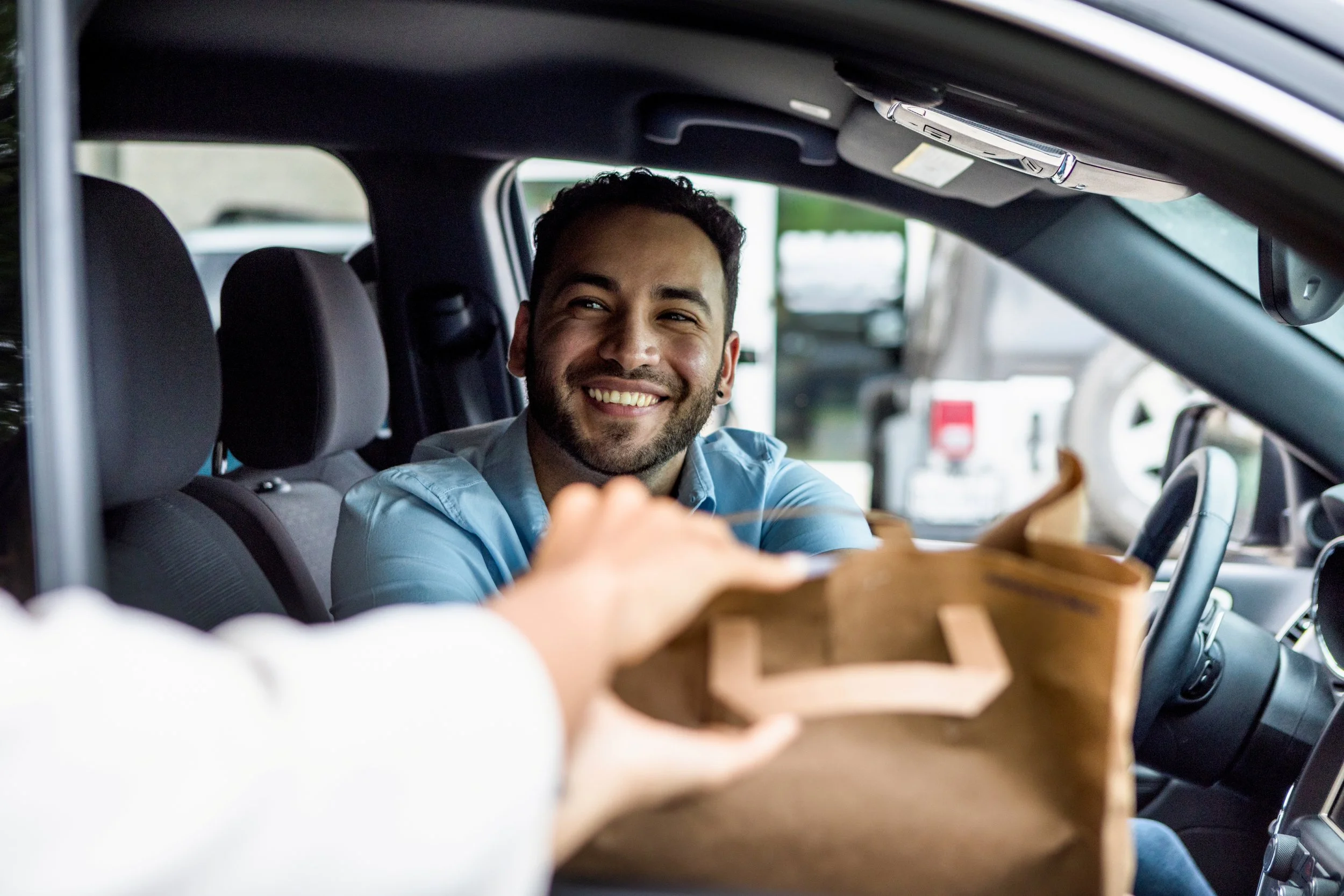 Smiling man in a car, reaching to receive a brown paper bag from someone outside the car.