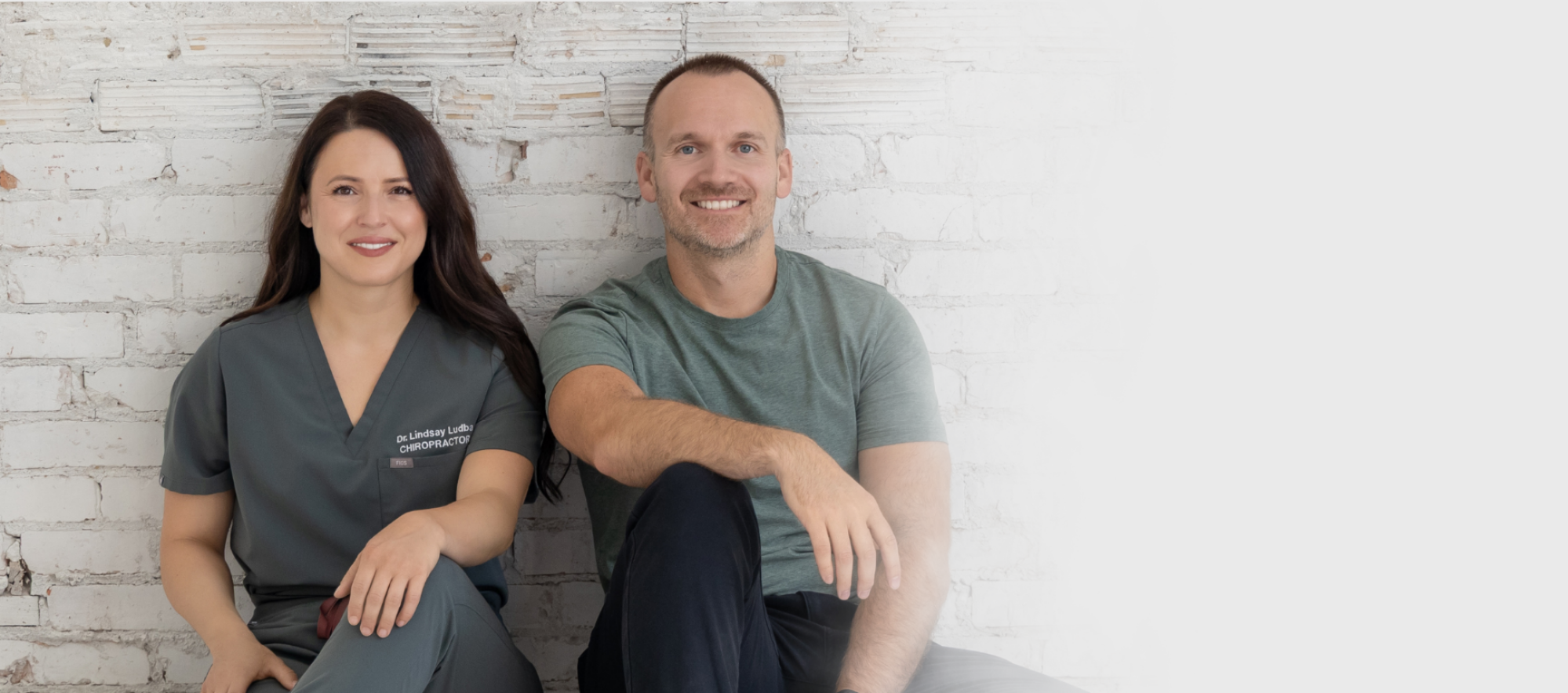 A woman in medical scrubs and a man in a casual t-shirt sitting against a white brick wall, smiling.