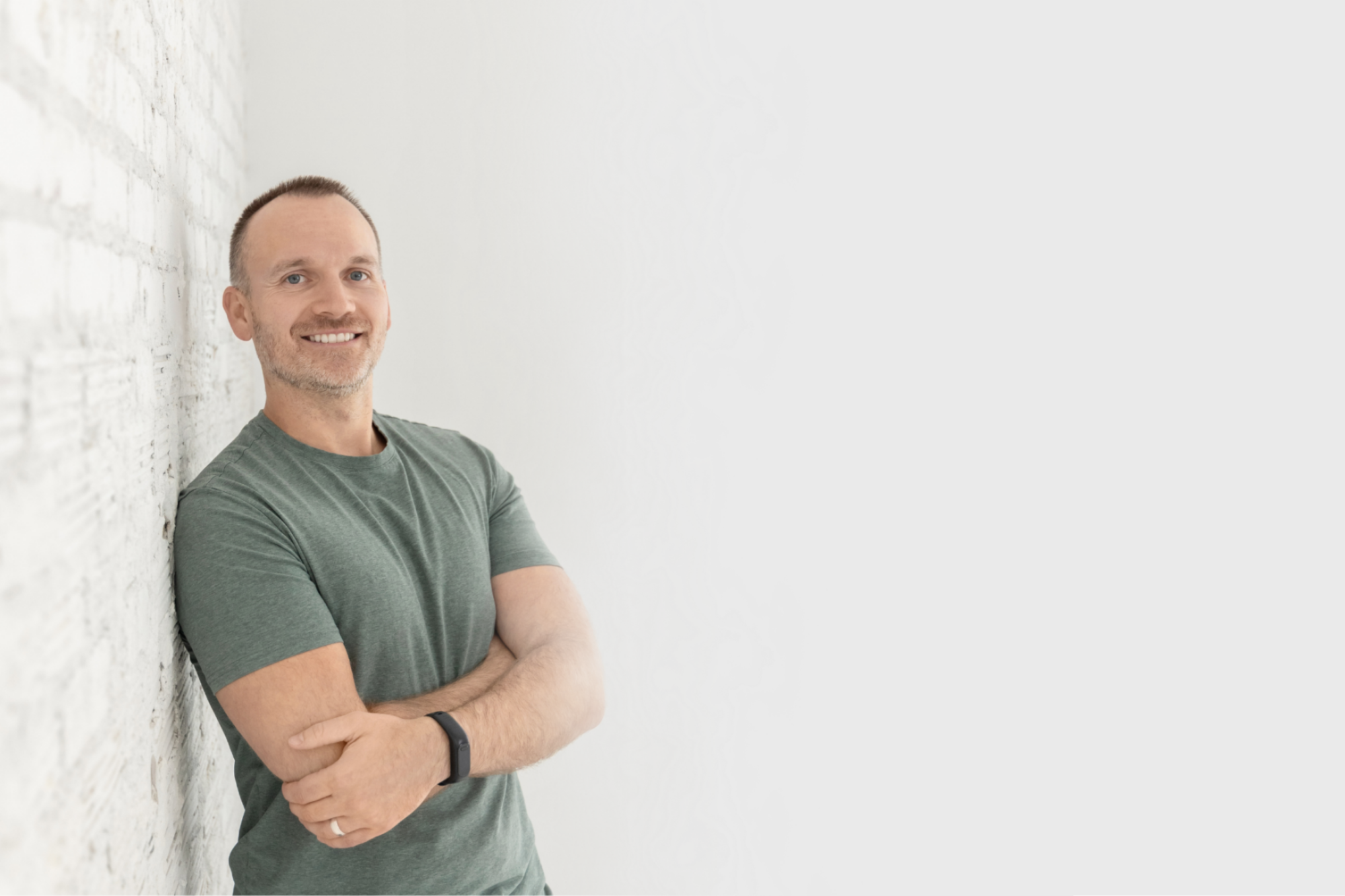 A smiling man with short hair and a beard wearing a green t-shirt, leaning against a white brick wall with arms crossed.