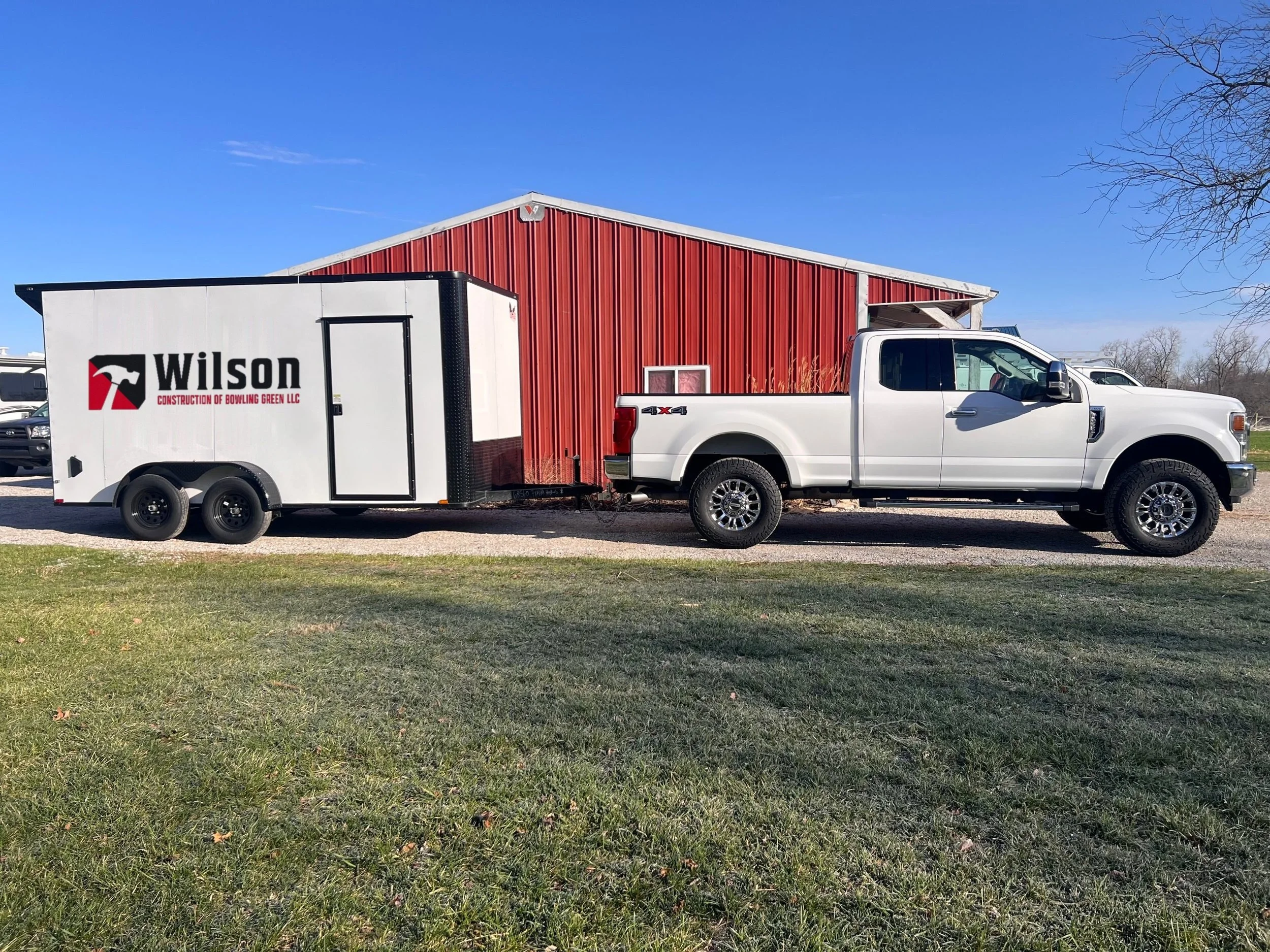 A white pickup truck with a trailer attached parked in front of a red barn on a sunny day with a clear blue sky.