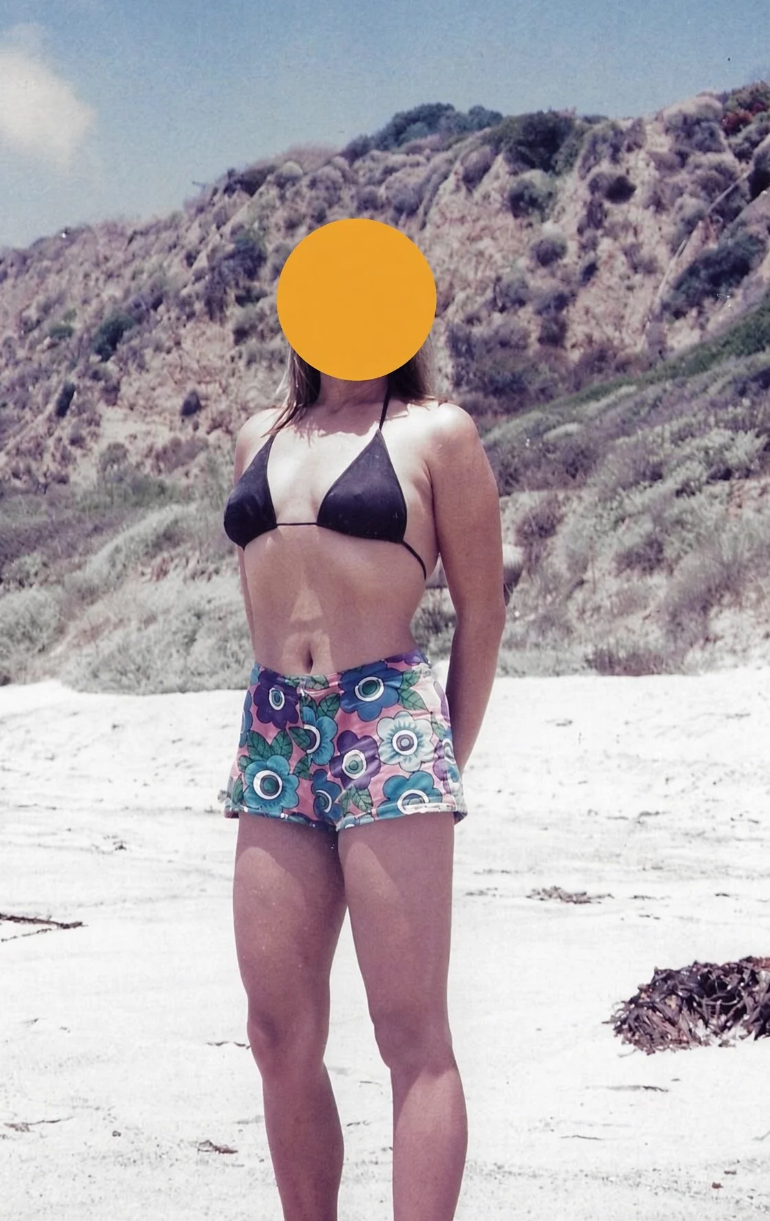 Beach portrait in a black bikini top and floral shorts, face gently obscured for privacy