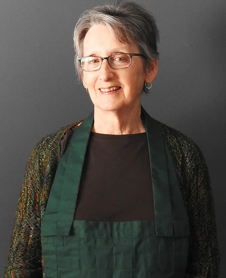 An older woman with short gray hair and glasses, smiling, wearing a black shirt, a multicolored sweater, and a dark green apron, standing against a dark gray background.