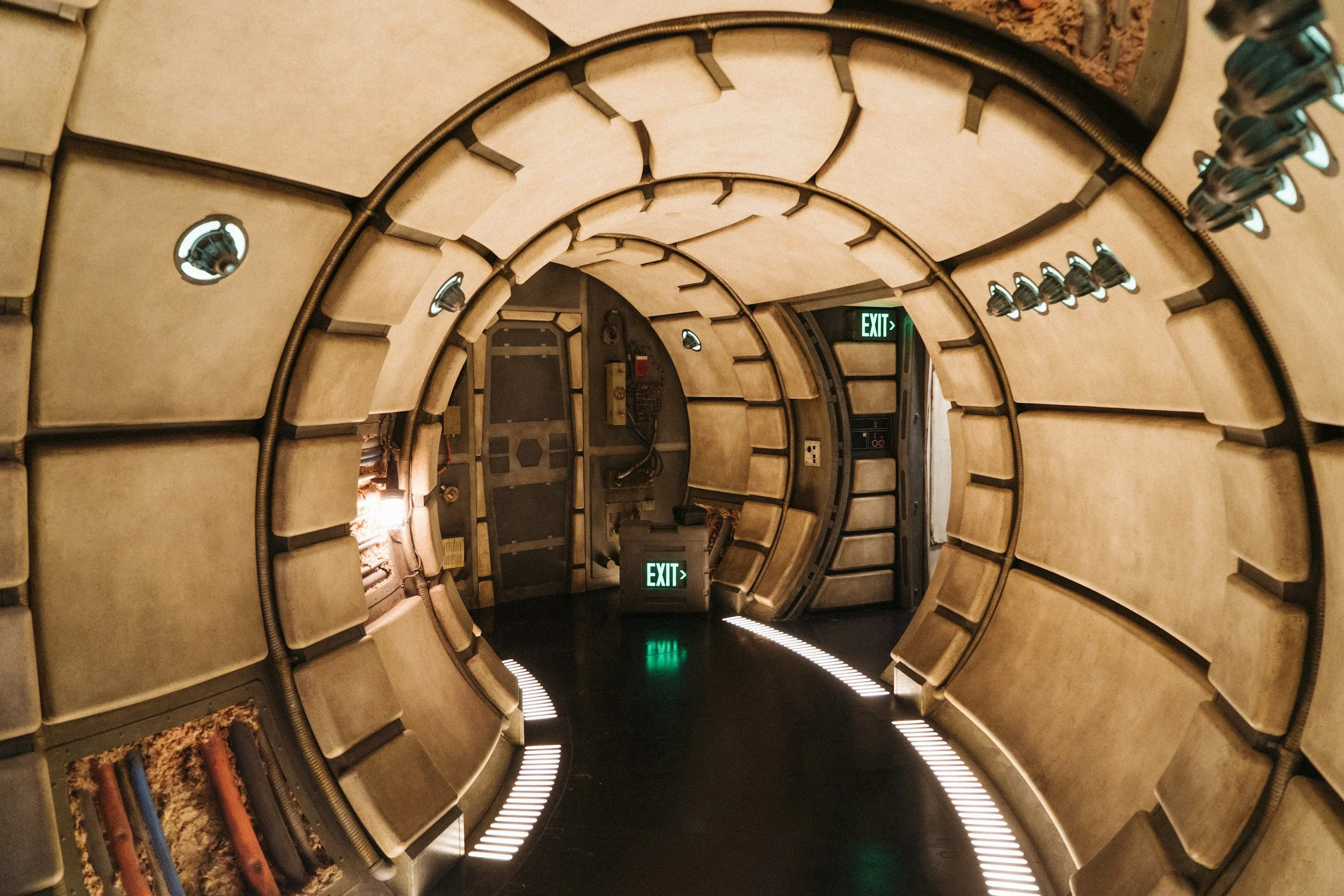 A futuristic, circular hallway with beige padded walls and an illuminated green exit sign at the end.