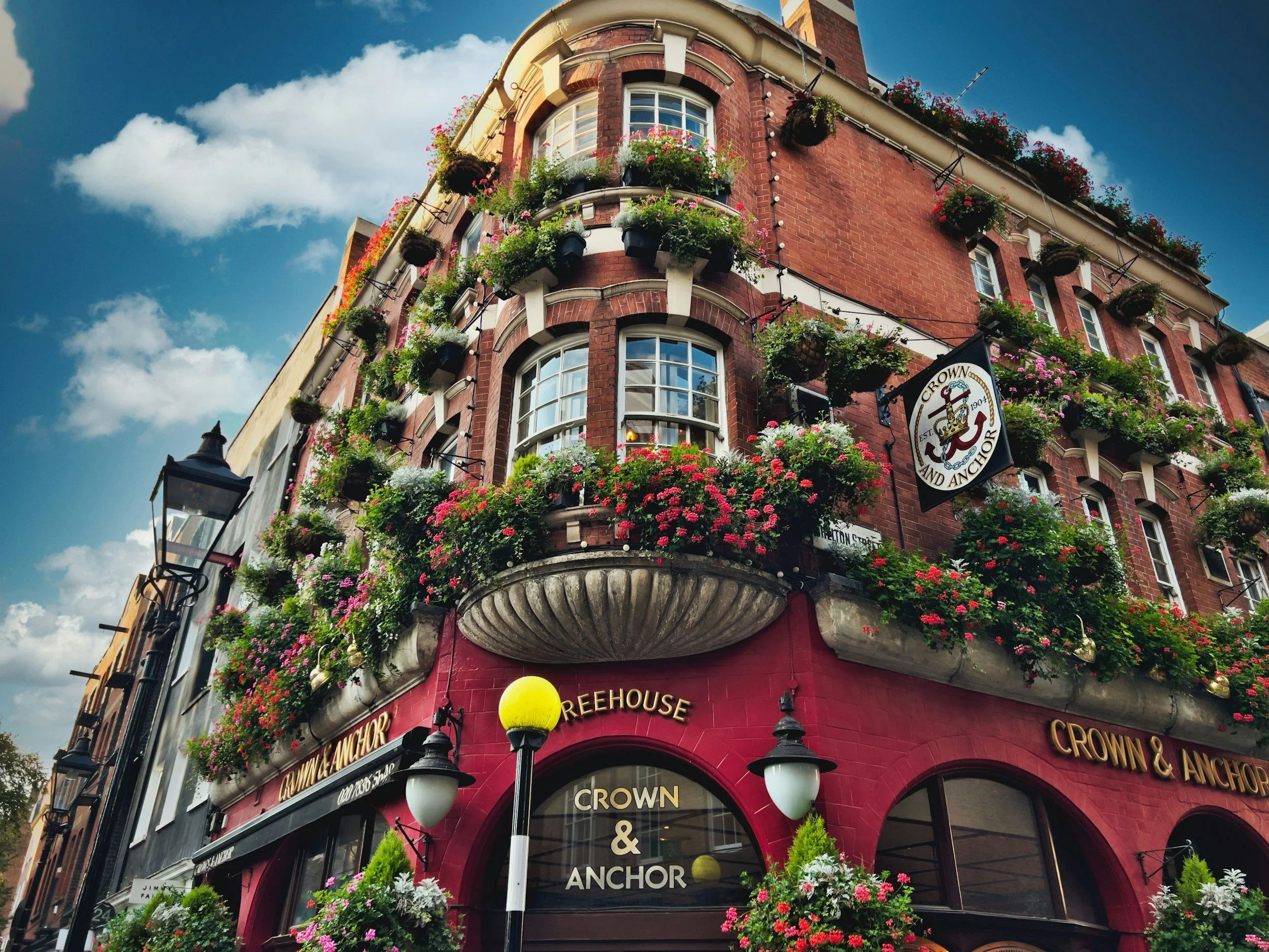 Upright red brick building with multiple floors and floral window boxes, featuring a sign for Crown & Anchor pub, and a street lamp in front, under a partly cloudy sky.