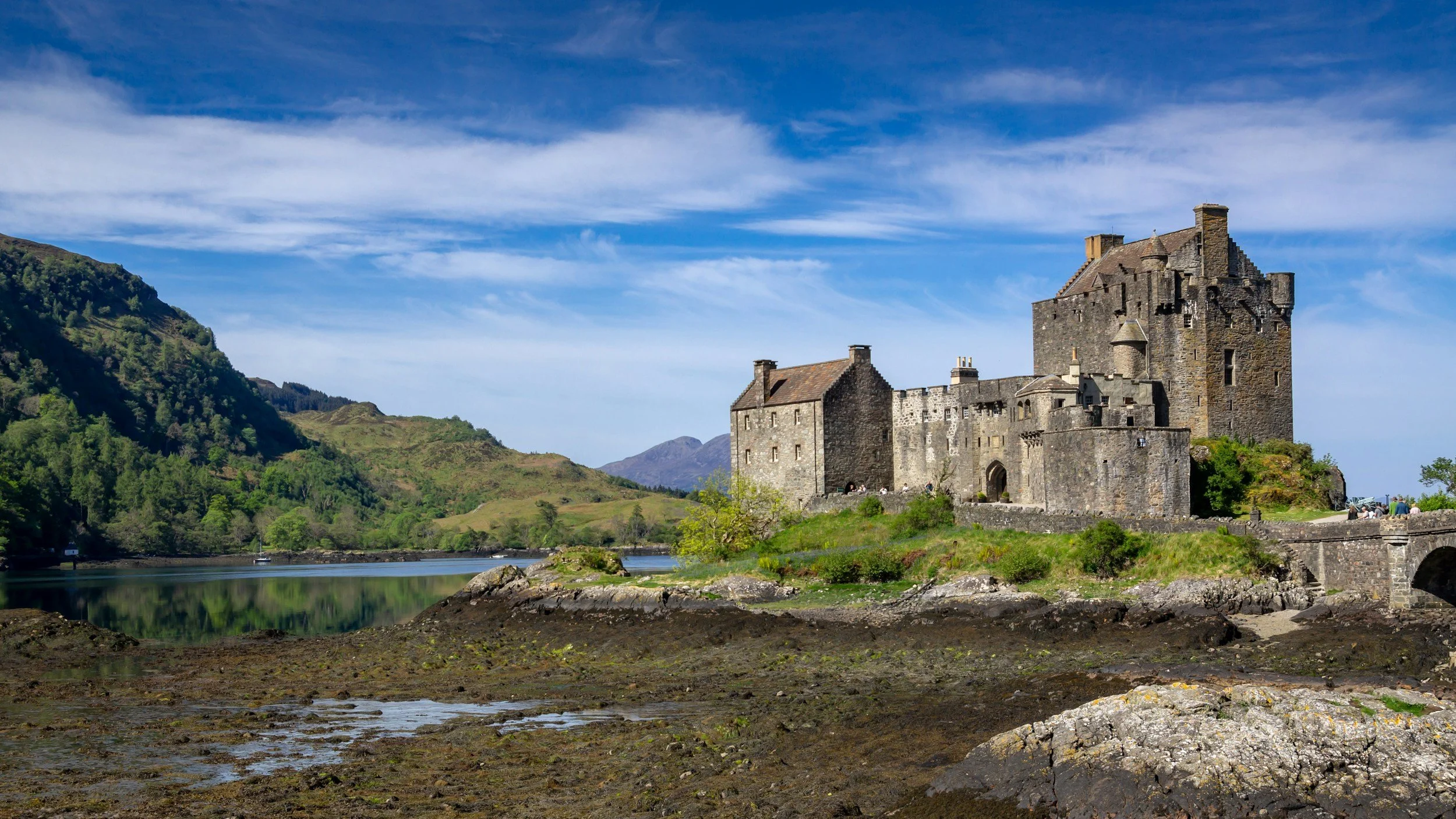 A historic stone castle located on a hillside near a body of water, with green trees and mountain scenery in the background under a partly cloudy sky.