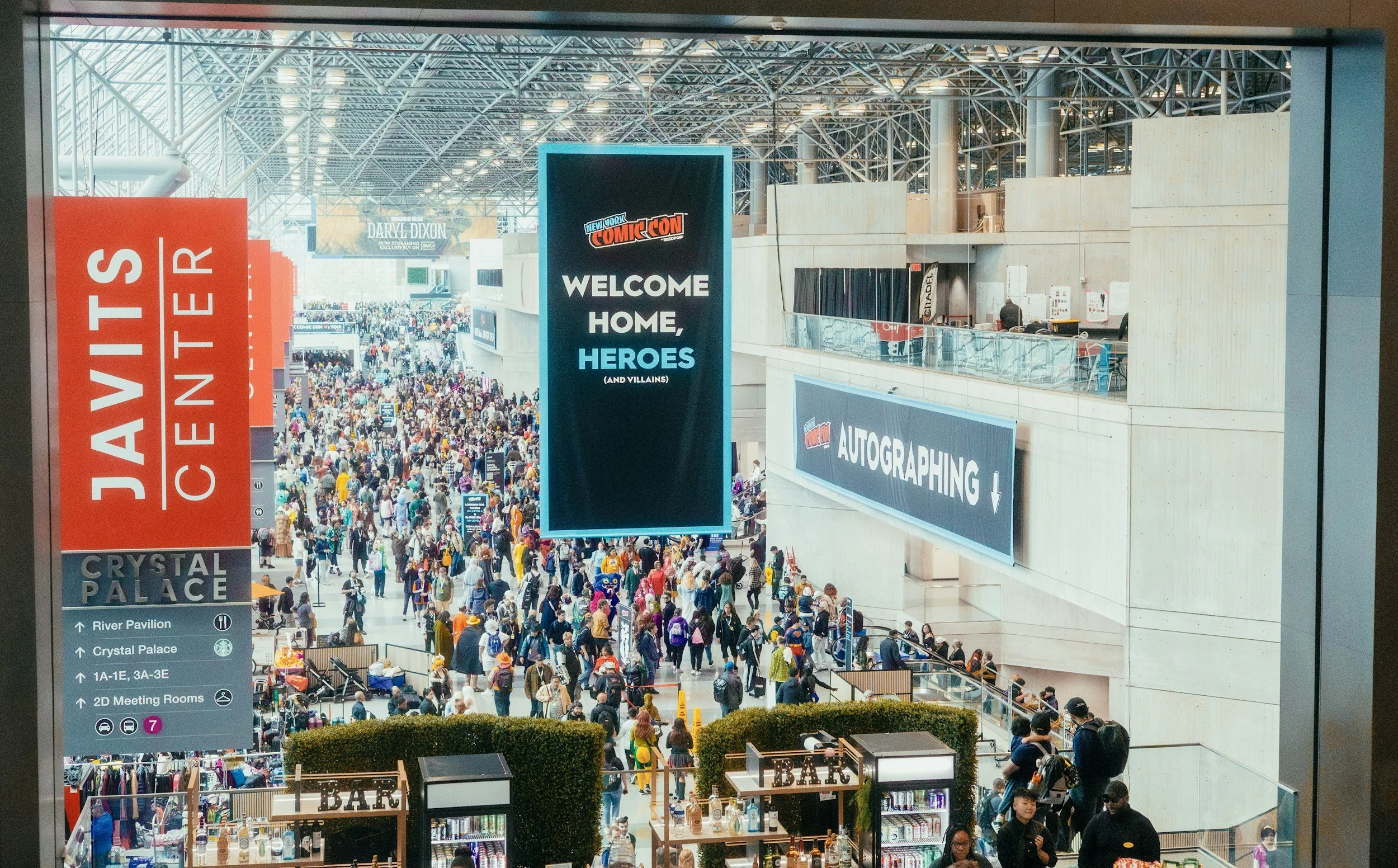 Crowd of people walking inside a convention center with large signs, including one saying 'WELCOME HOME, HEROES' and another for 'JAVITS CENTER.' The ceiling is high with exposed steel beams and bright lighting. There are souvenir and food booths, some with 'BAR' signs, and informational directional signs.