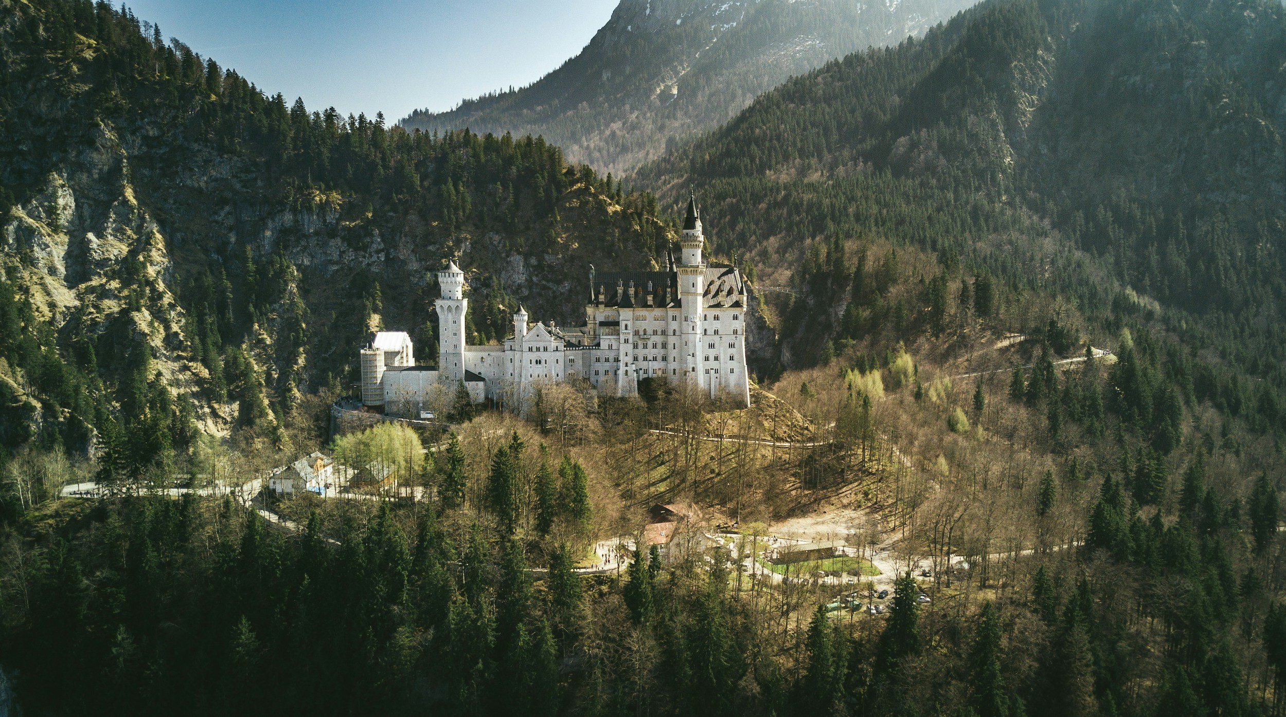 A white castle with tall towers and spires situated on a hill, surrounded by dense evergreen trees and mountain terrain in the background.