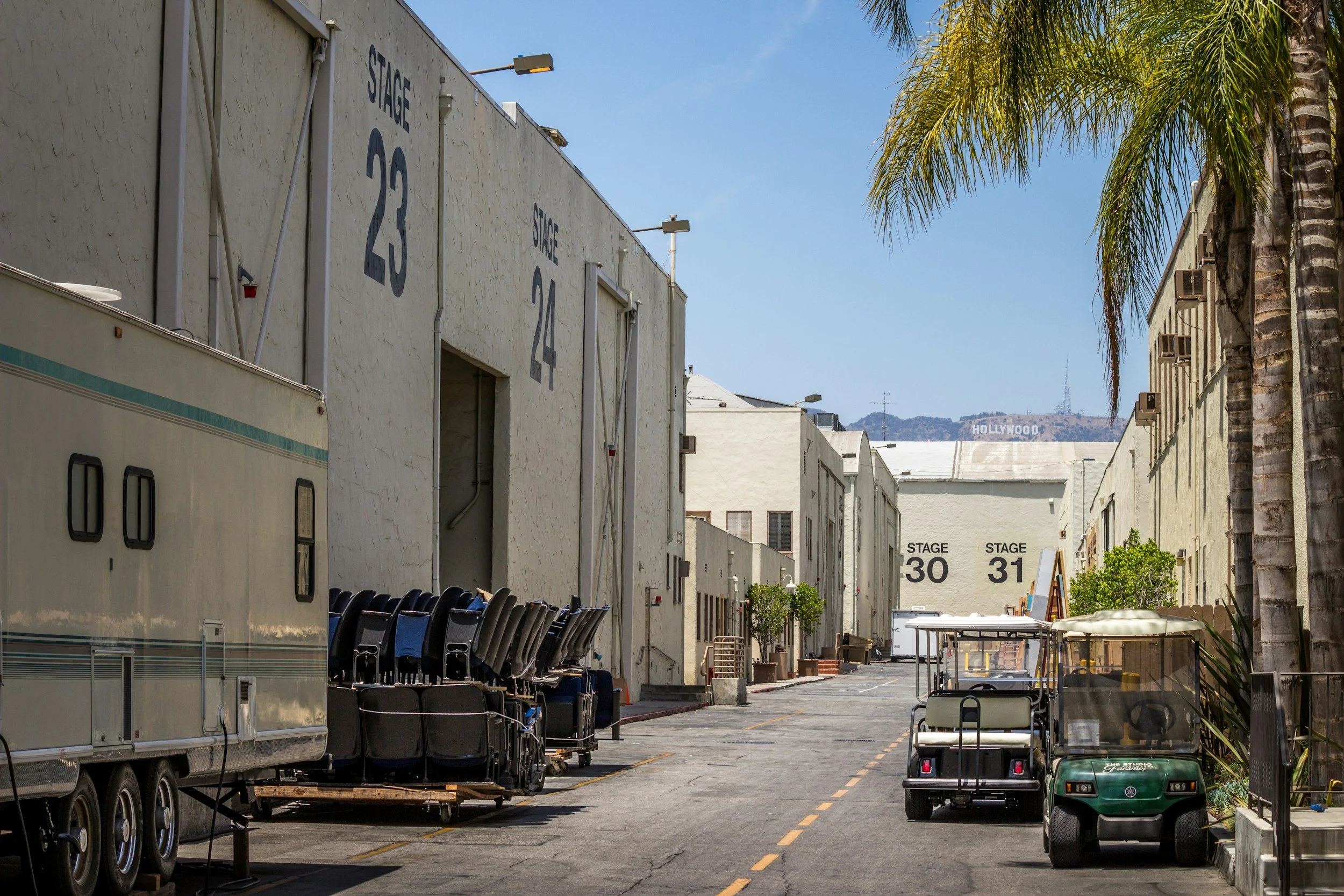 Empty street backstage area with white buildings labeled 'Stage 30' and 'Stage 31', palm trees on the right, trailers, golf carts, and chairs lined up.
