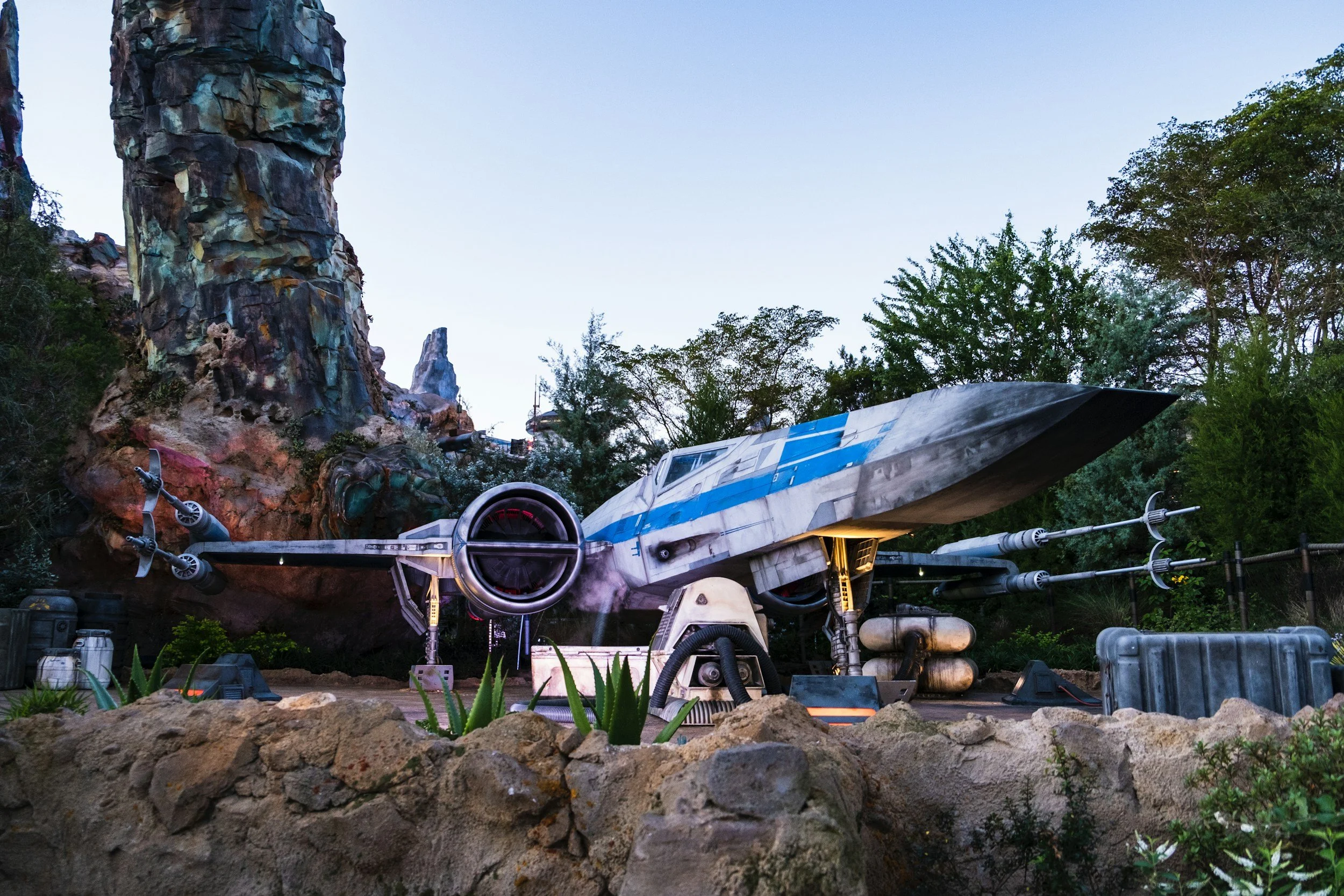 A detailed miniature model of the x-wing spaceship from Star Wars, with a rocky landscape and greenery around it, and a tall rock formation in the background.