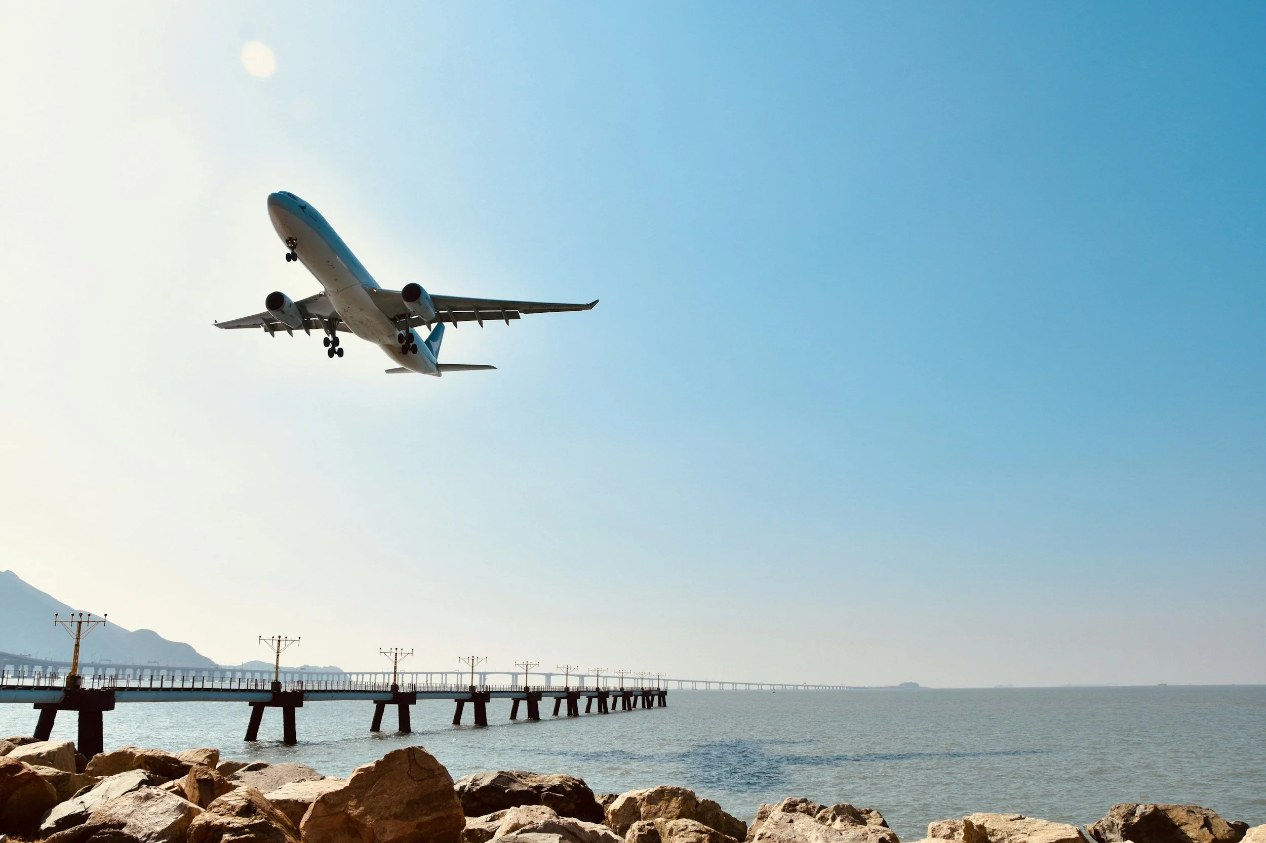 An airplane flying above a coastal pier with rocks in the foreground and a mountain in the background.