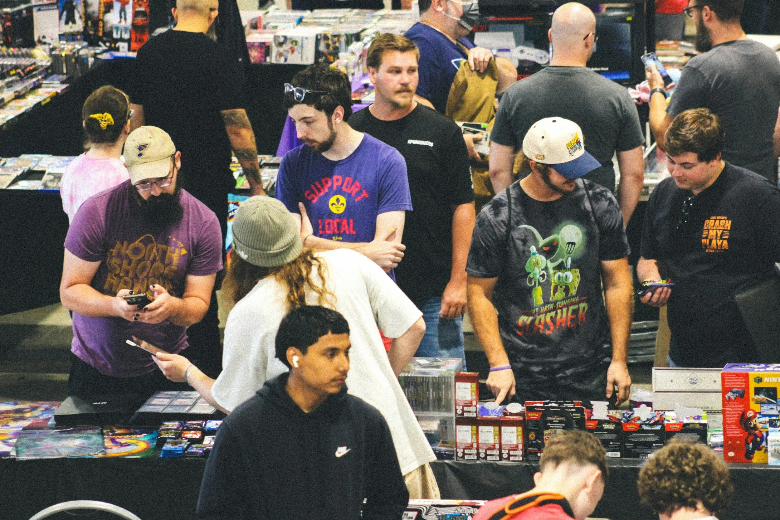 People browsing and purchasing items at a convention or comic book store, with tables displaying collectibles and comics, some focused on gaming and pop culture.