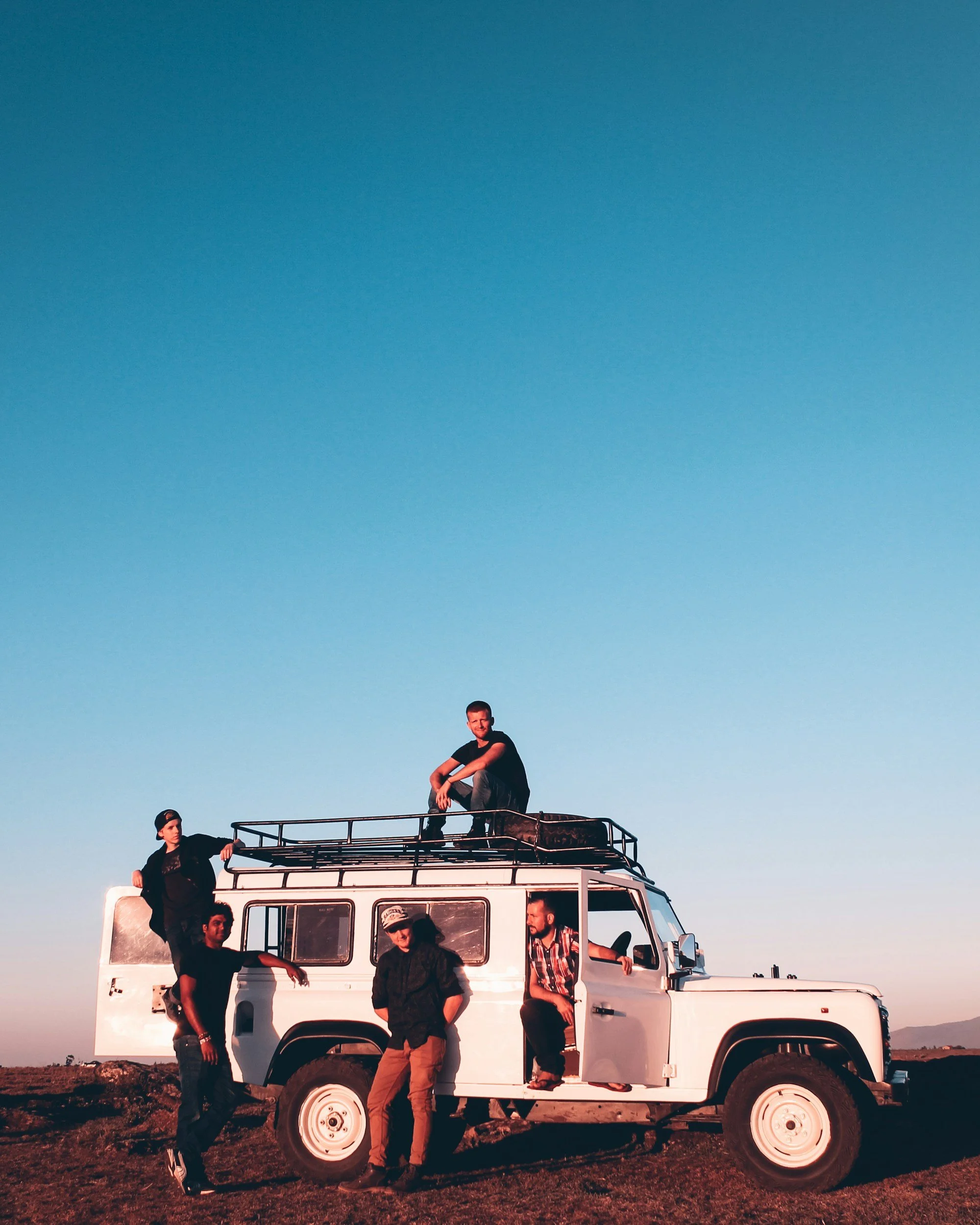 Six men gathered around and on a white off-road vehicle with a roof rack, in an open field at sunset, with a clear sky in the background.