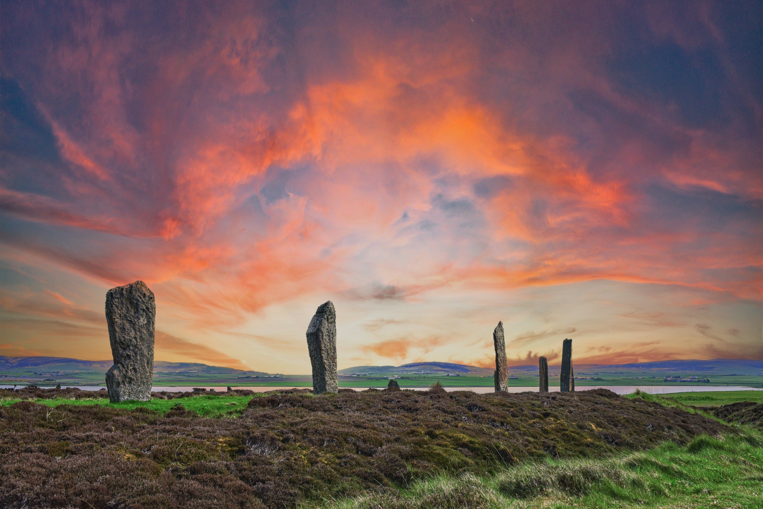 A landscape with five standing stones of varying sizes in a grassy and moor-like area, with dark mountain ranges in the background and a colorful sunset sky with pink and orange clouds.