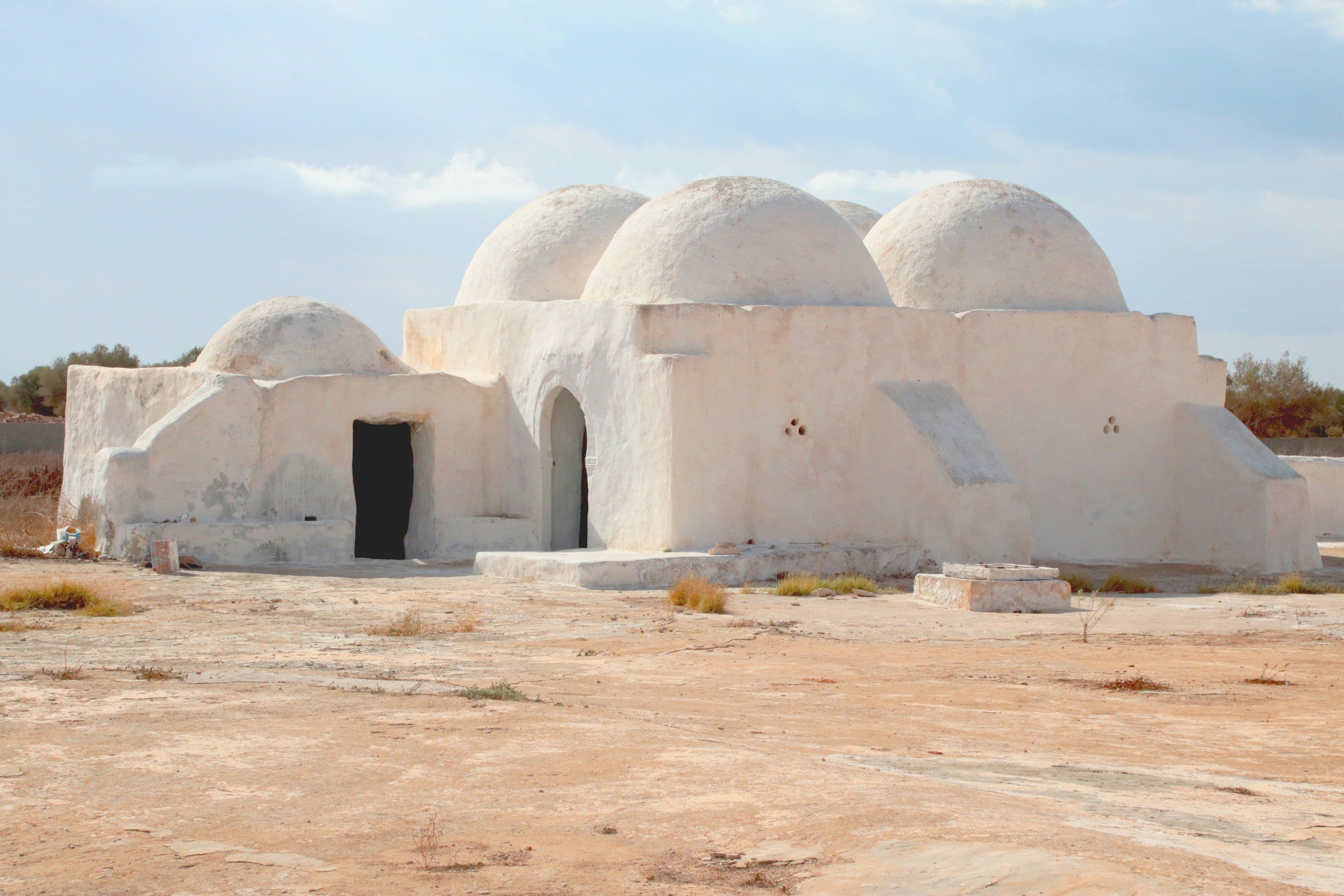 White adobe building with rounded domes on top, set in a desert landscape with sparse vegetation and a cloudy sky.