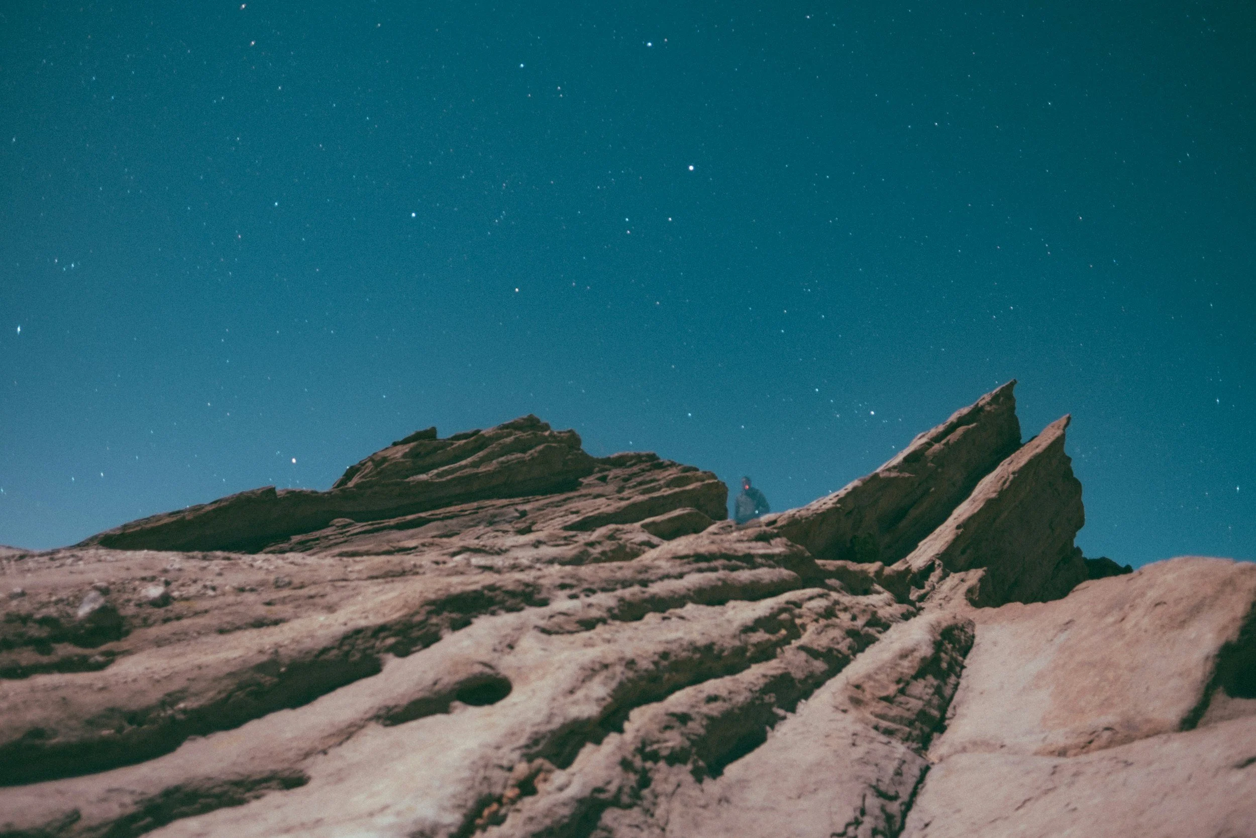 Night sky with numerous stars above rocky desert landscape, including a person sitting on the rocks.