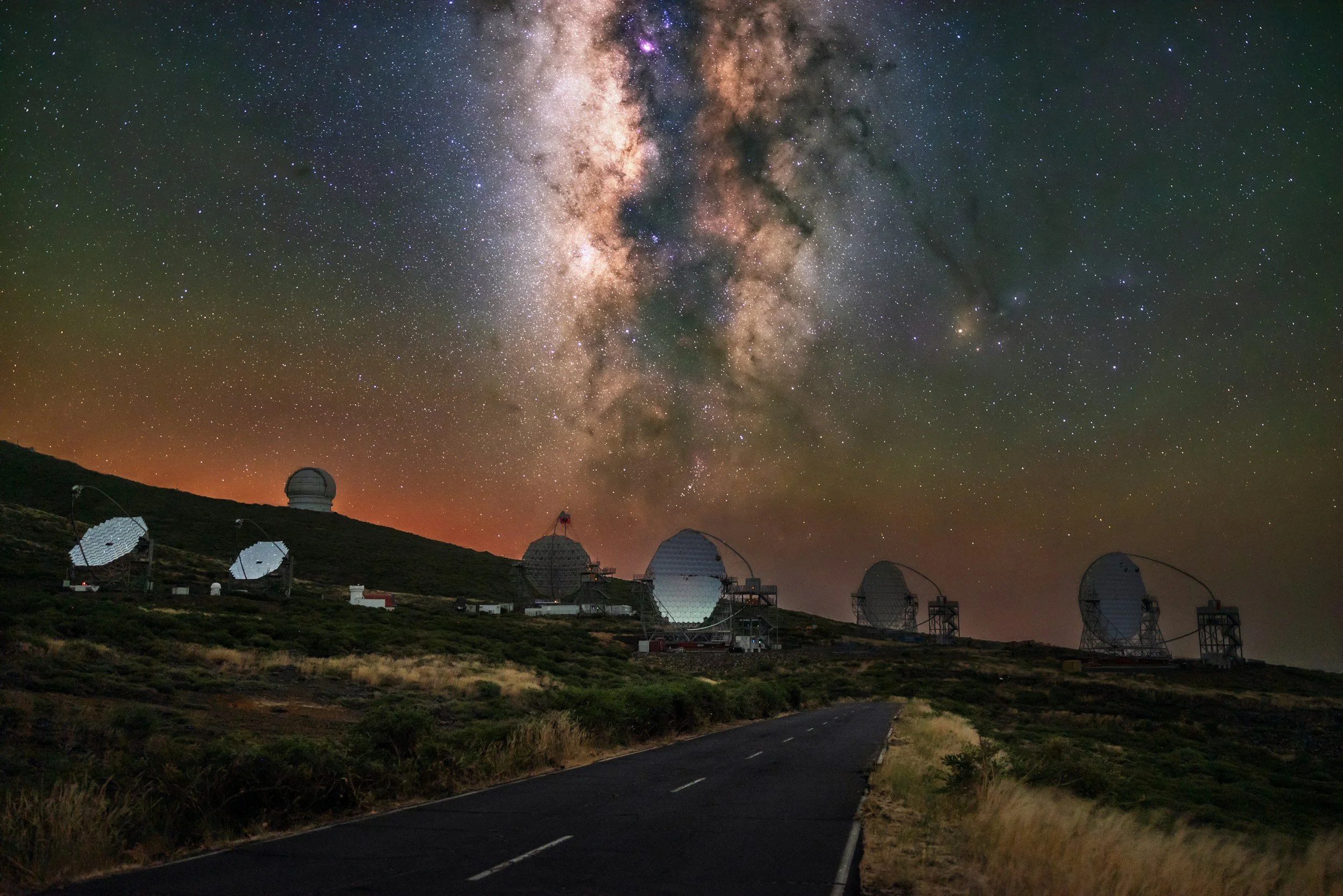 Night sky with the Milky Way galaxy visible above a landscape with telescopes and a winding road.