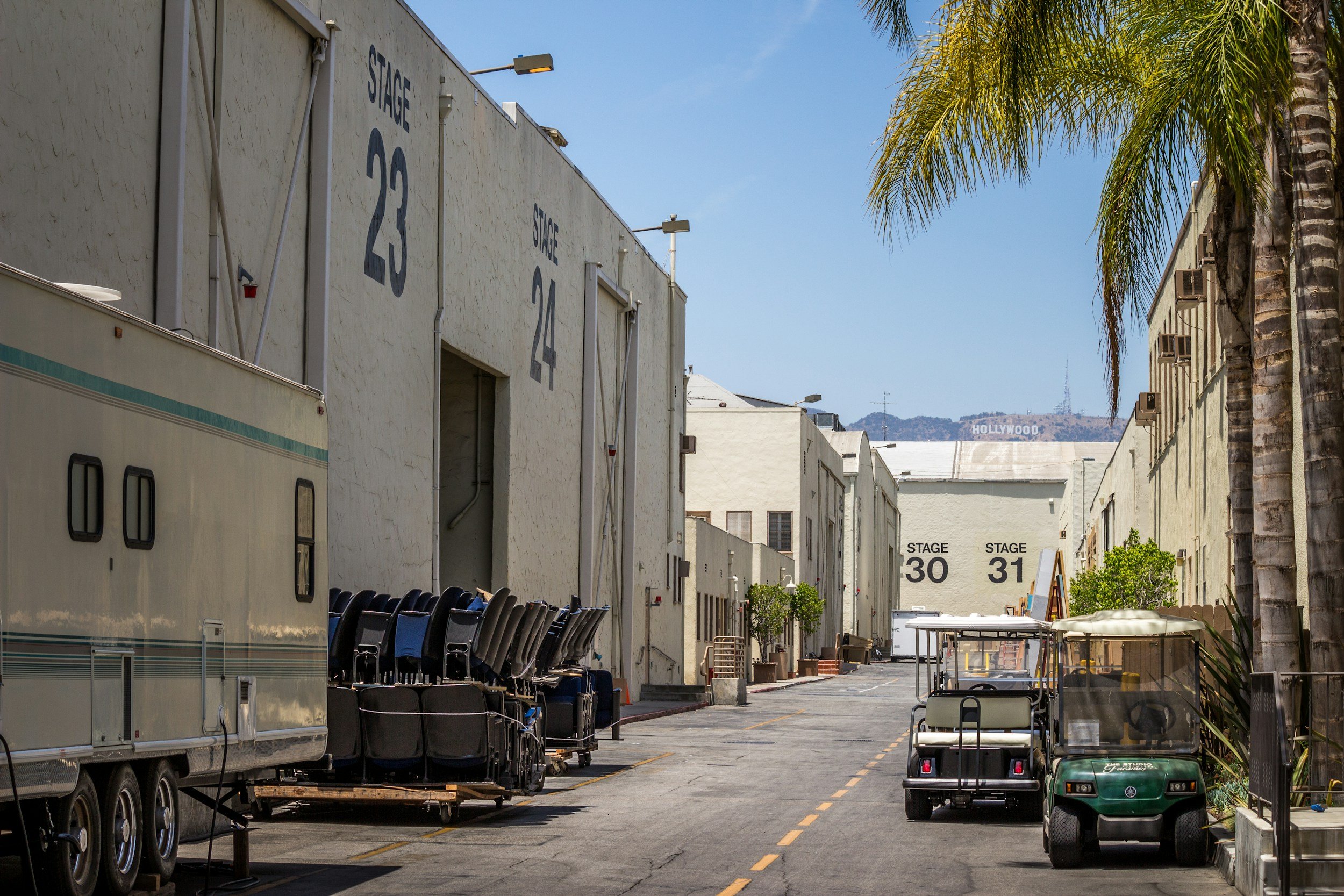 Empty outdoor alley with white buildings labeled with stage numbers, palm trees, parked golf carts, and a sign for Hollywood in the distance.