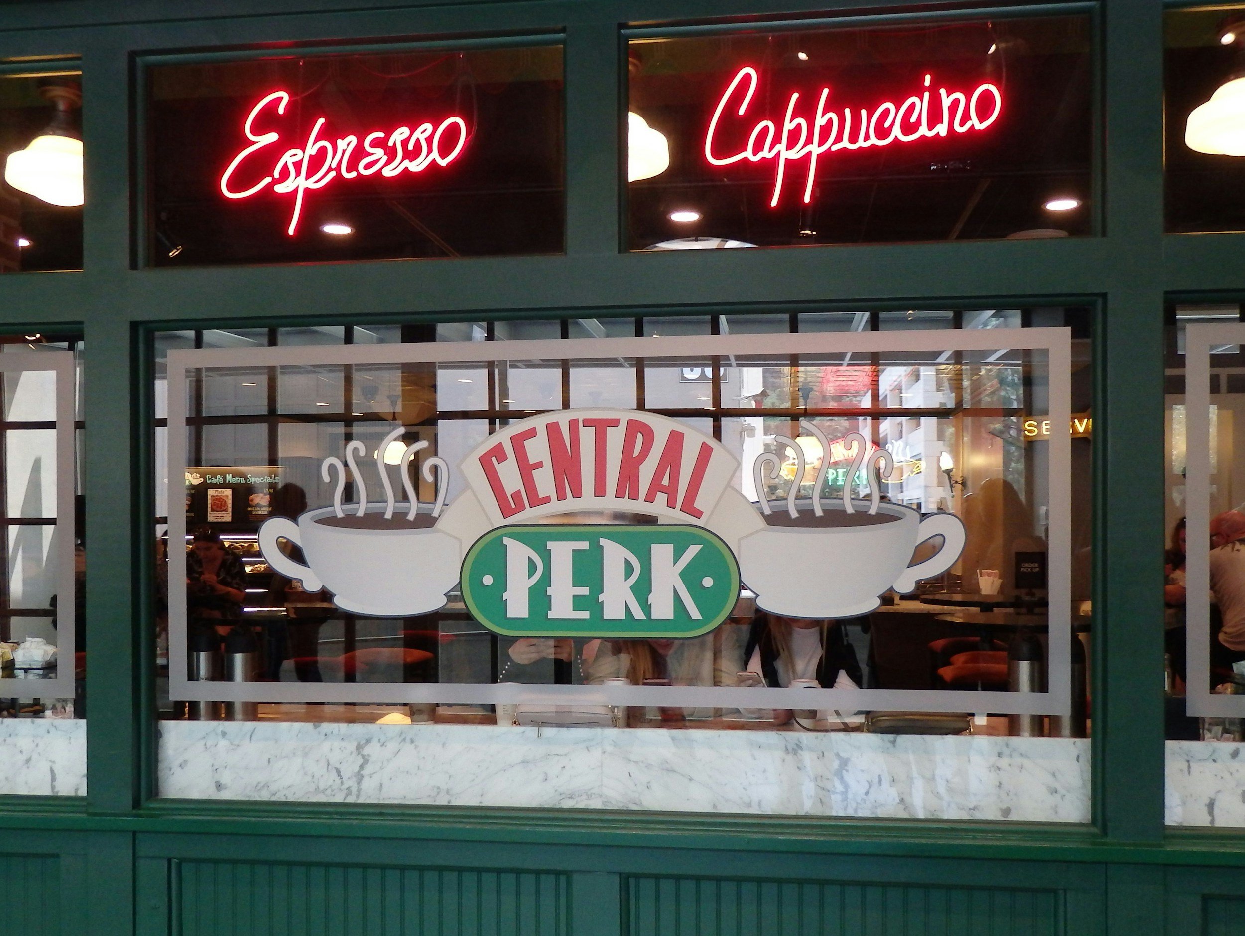 Neon signs indicating Espresso and Cappuccino above the window of a cafe, with people visible through the window inside.