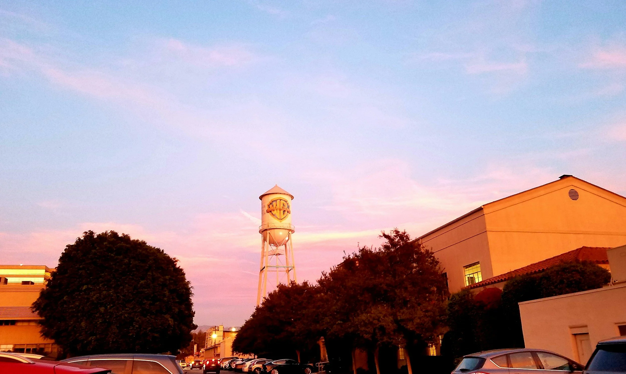 Sunset sky with pink and purple hues over a building and trees, with a water tower and parked cars in the foreground.