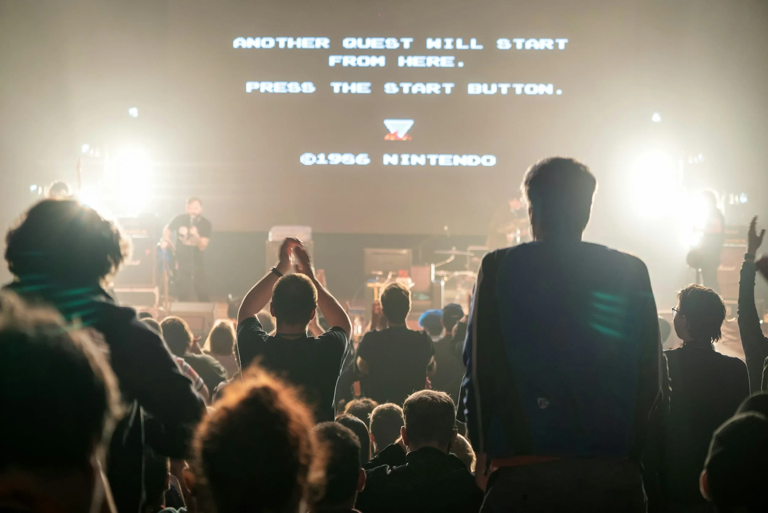 Crowd of people attending a concert or event, facing a stage with bright lights and a large screen displaying instructions to start from another quest hill, with the Nintendo logo and copyright date 1986.