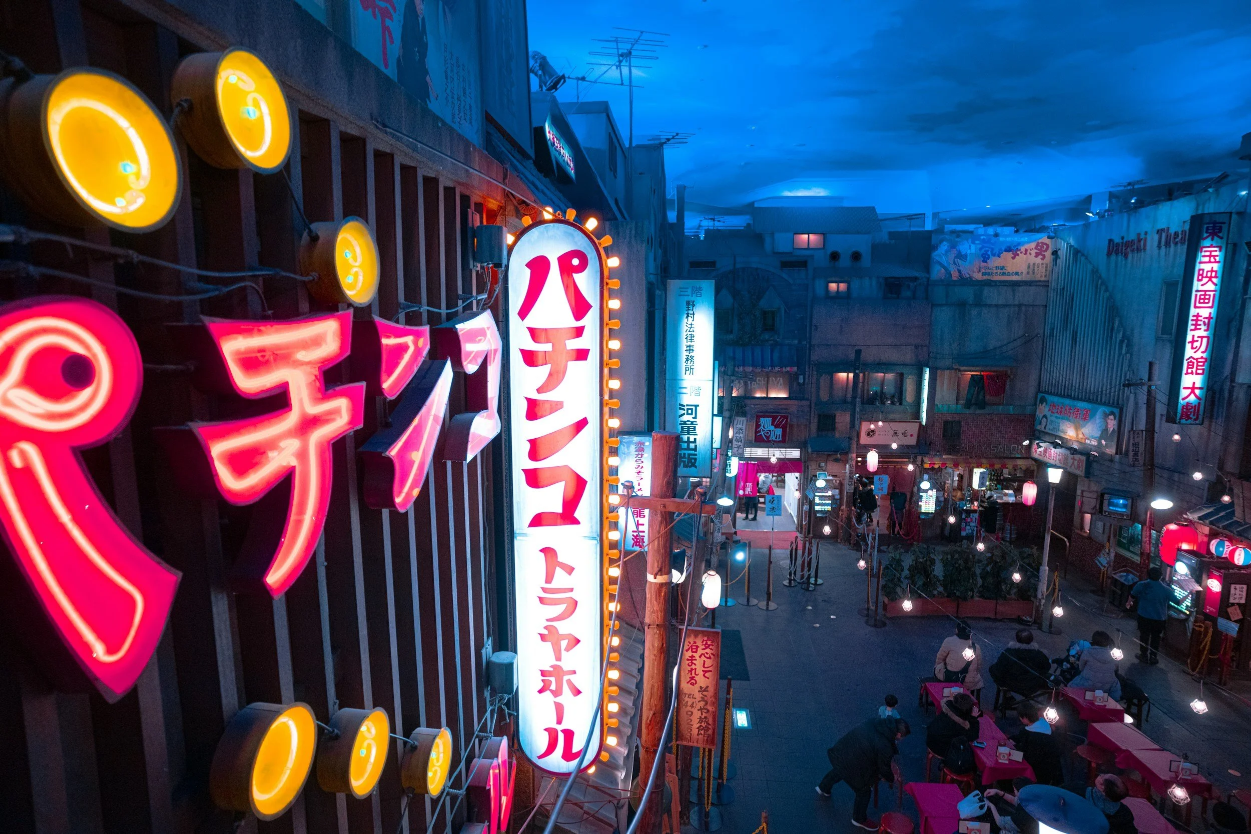 Night scene of a busy street with colorful neon signs in Japanese, illuminated lanterns, and people walking and dining outside.