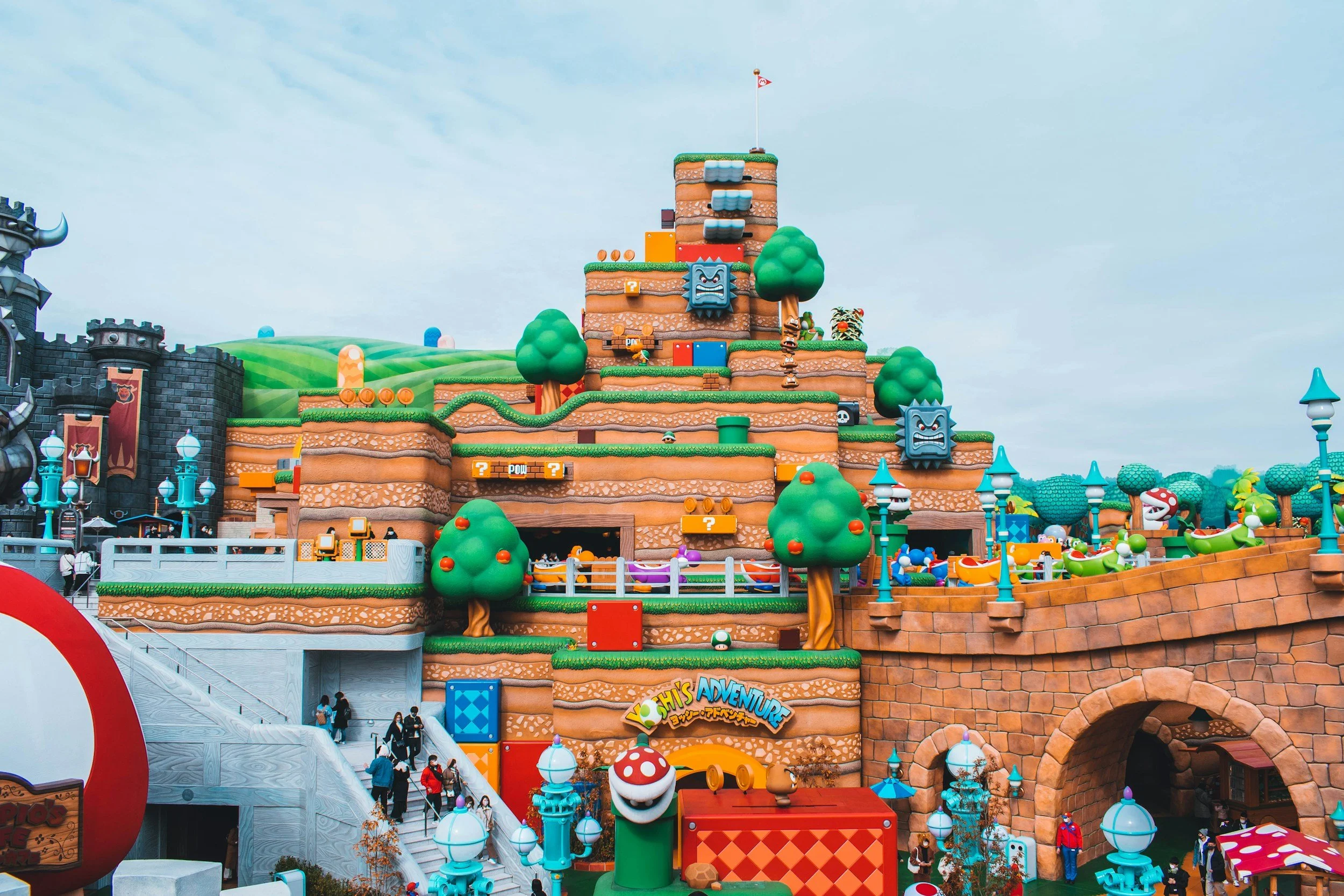 Colorful Mario-themed amusement park with structures resembling brick blocks, pipes, and brick walls, and decorations like green trees, mushrooms, and Thwomps from the Super Mario game.